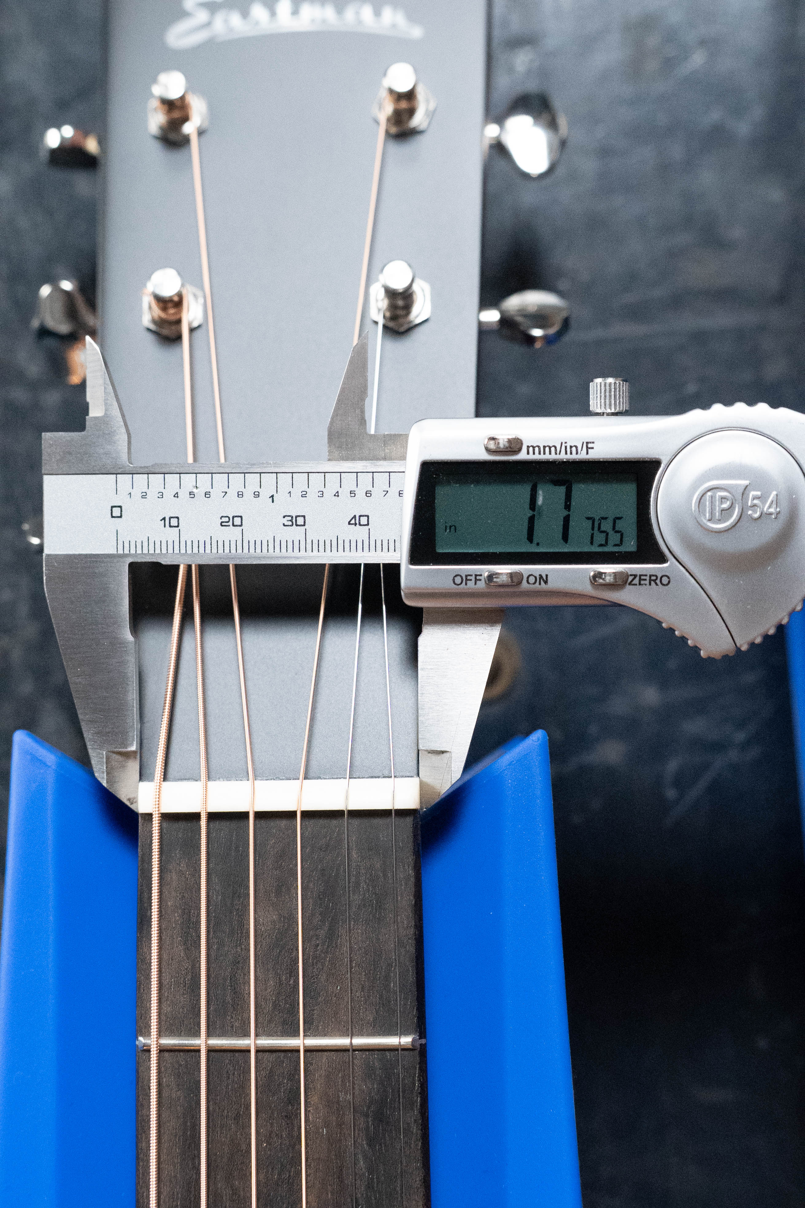 A digital caliper measures the width at the nut of an Eastman Guitars E2D-DLX-SB Cedar/Sapele Sunburst Dreadnought w/Fishman Pickup #1532, displaying 1.7 inches. The guitar stands upright in a blue stand on a dark surface.