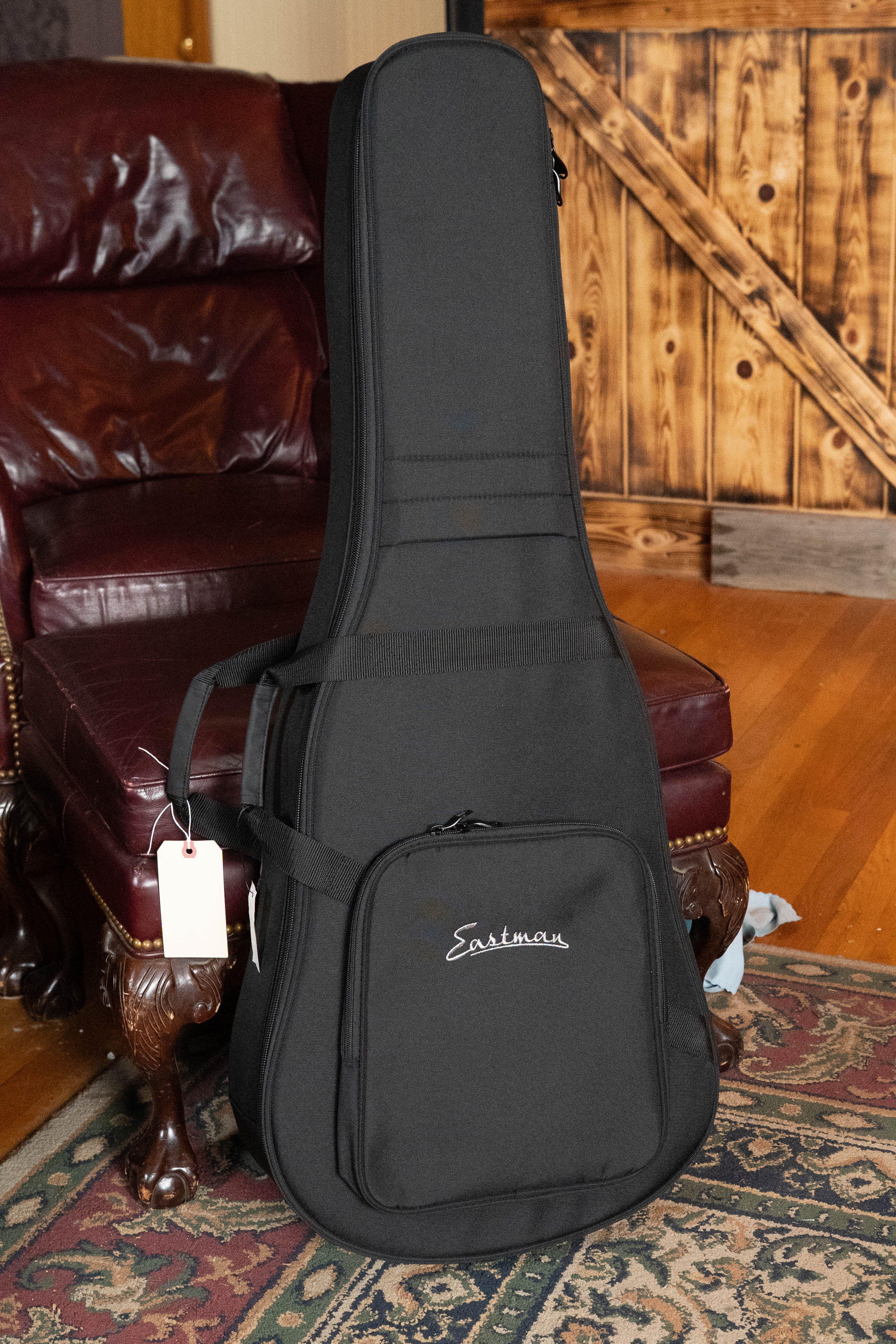 A black Eastman Guitars case with front pocket and name tag stands upright on a vintage red leather chair, ready to protect your Eastman E2D-DLX-SB Cedar/Sapele Sunburst Dreadnought Guitar w/Fishman Pickup #1532.