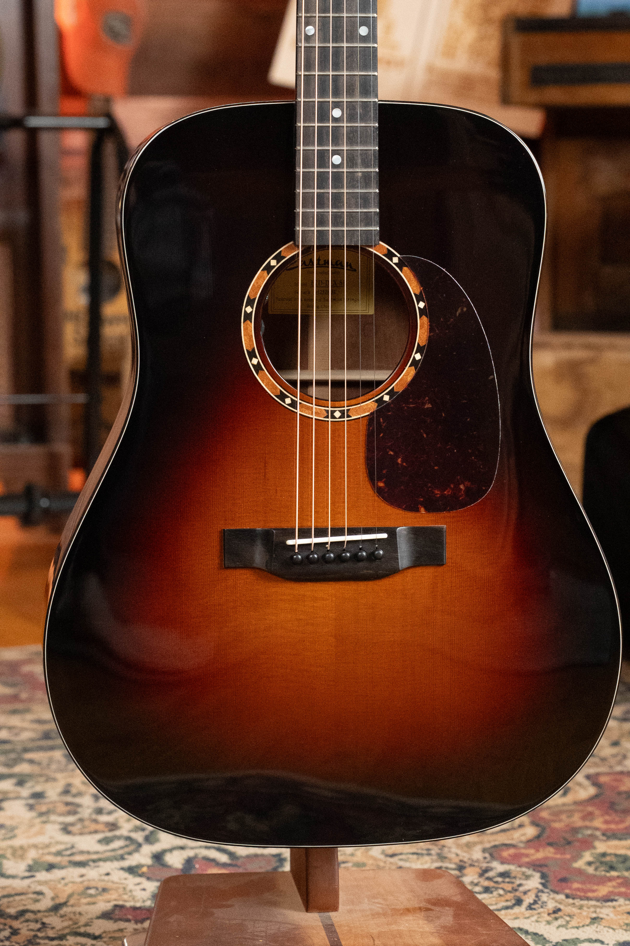 A close-up of the Eastman Guitars E2D-DLX-SB Cedar/Sapele Sunburst Dreadnought w/Fishman Pickup (#1532), featuring pearl inlays around the sound hole and a tortoiseshell pickguard, shown on an indoor stand.