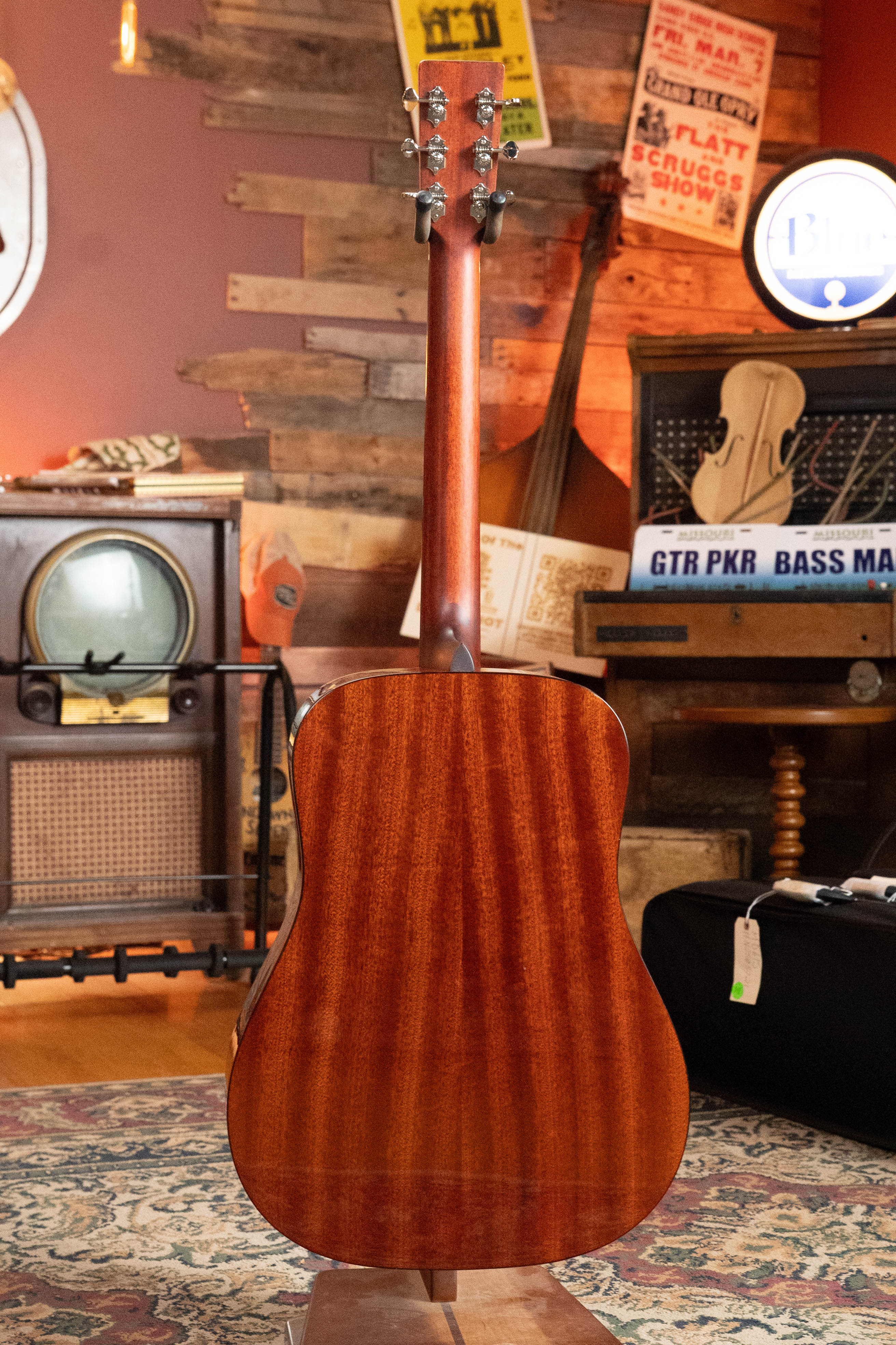 The Eastman Guitars E2D-DLX-SB Cedar/Sapele Sunburst Dreadnought Guitar w/Fishman Pickup #1532 stands upright with its back showing in a cozy, vintage room adorned with musical instruments, retro signs, and patterned rugs.