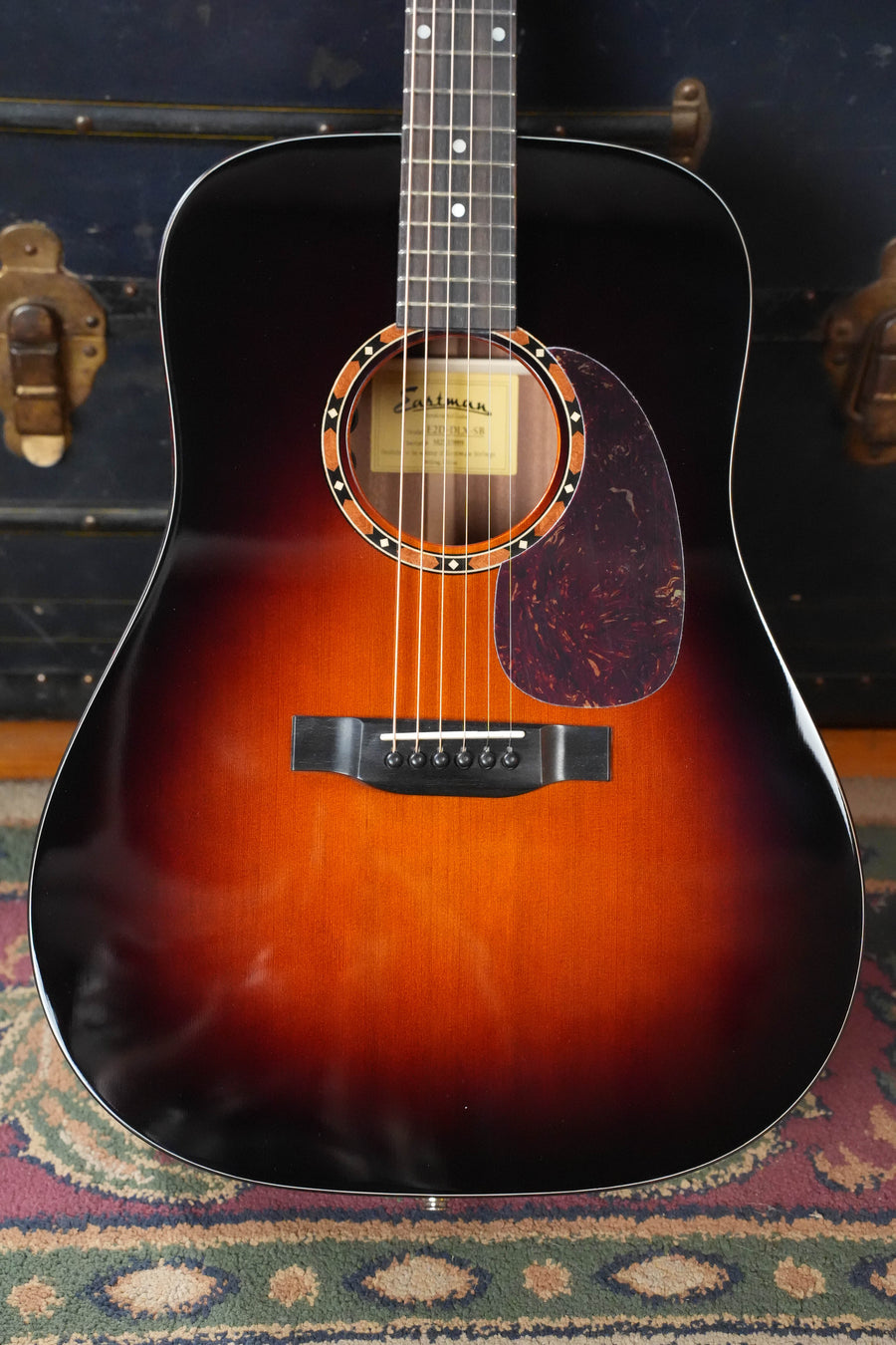 An Eastman Guitars E2D-DLX-SB Cedar/Sapele Sunburst Dreadnought w/Fishman Pickup (#5889) stands upright on a patterned rug, featuring a tortoiseshell pickguard and set before a vintage trunk with brass hardware.