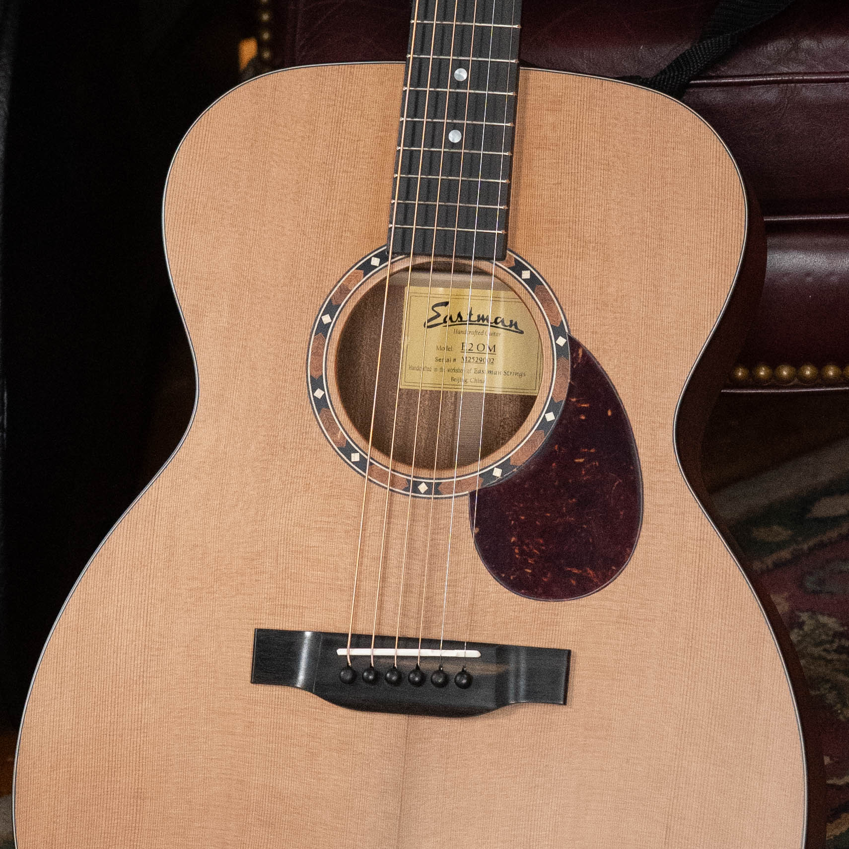 A close-up of an Eastman Guitars E2OM Cedar/Sapele Orchestra Model Acoustic Guitar #9002 highlights its strings, soundhole, solid cedar top, and pickguard, with a leather chair and patterned rug in the background.