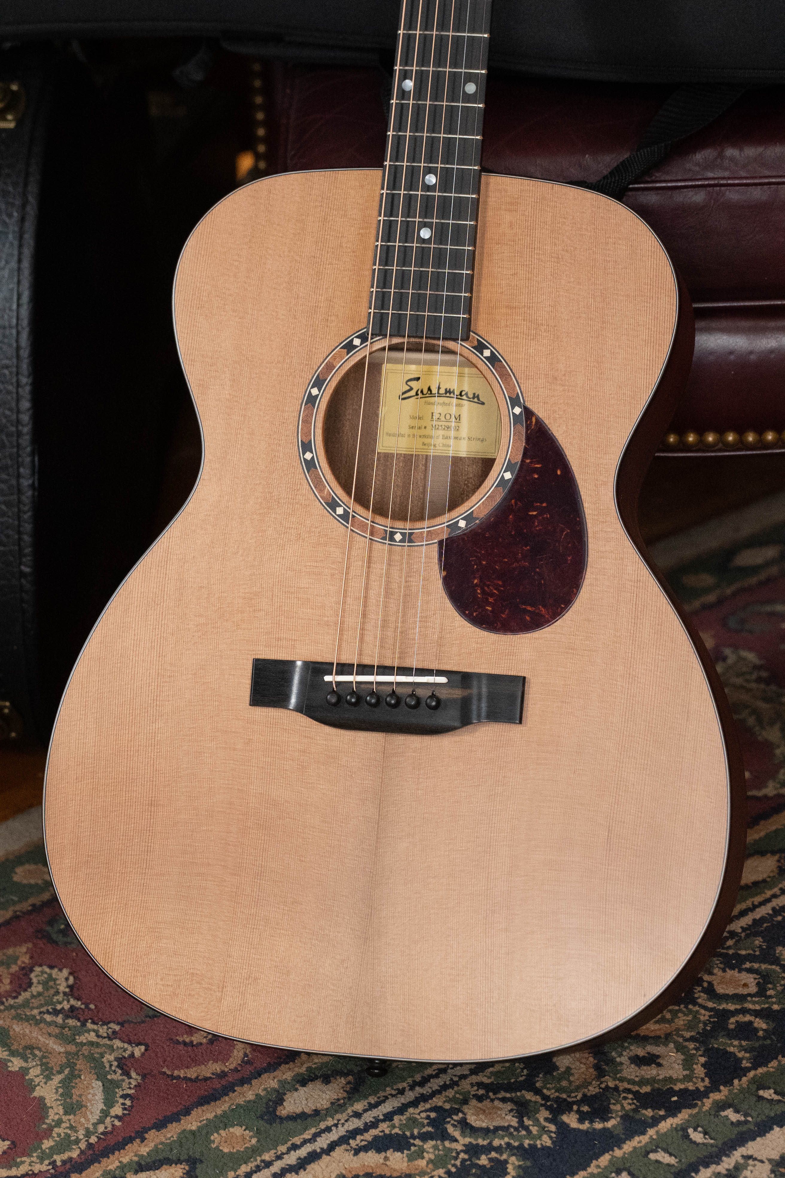 A close-up of the Eastman Guitars E2OM Cedar/Sapele Orchestra Model Acoustic Guitar #9002, featuring a solid cedar top, natural finish, dark pickguard, and six strings, resting on a patterned rug with the Eastman label visible inside the sound hole.