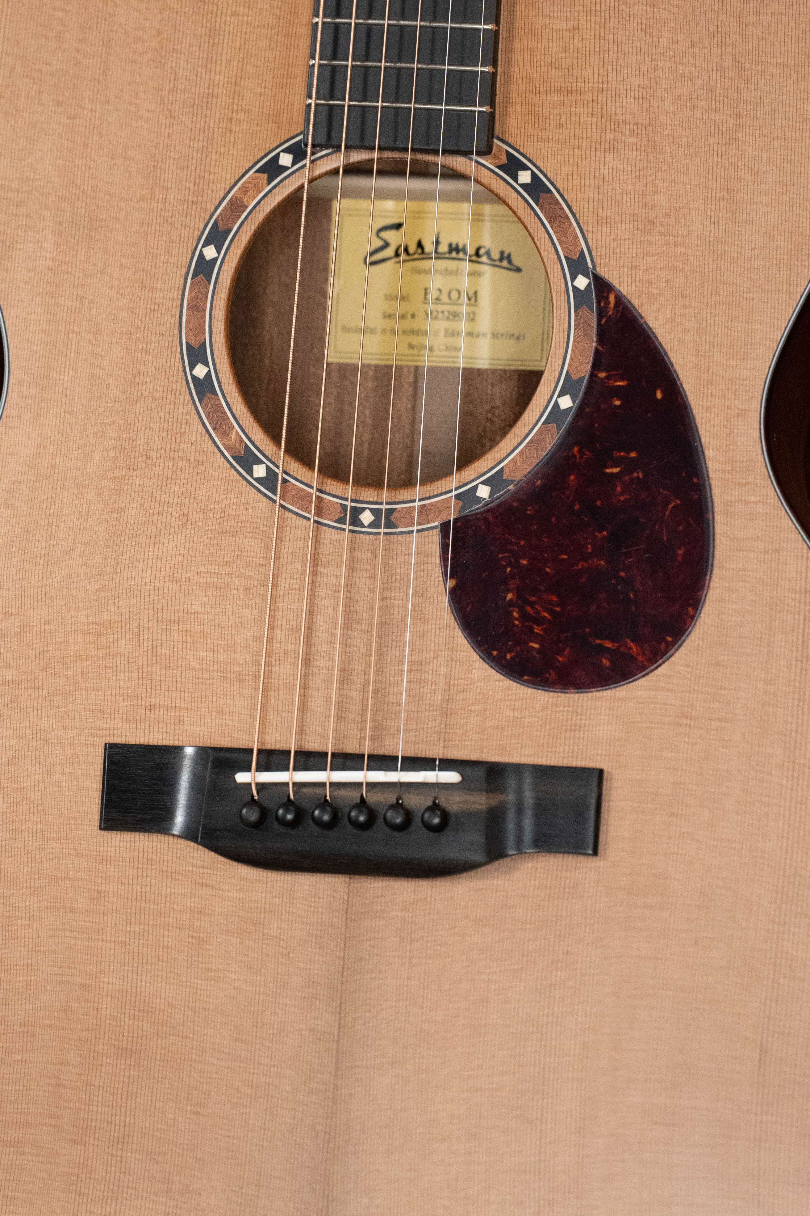 Close-up of the Eastman Guitars E2OM Cedar/Sapele Orchestra Model Acoustic Guitar #9002, highlighting its solid cedar top, decorative soundhole rosette, inner label, dark pickguard, bridge, and strings on a natural finish.