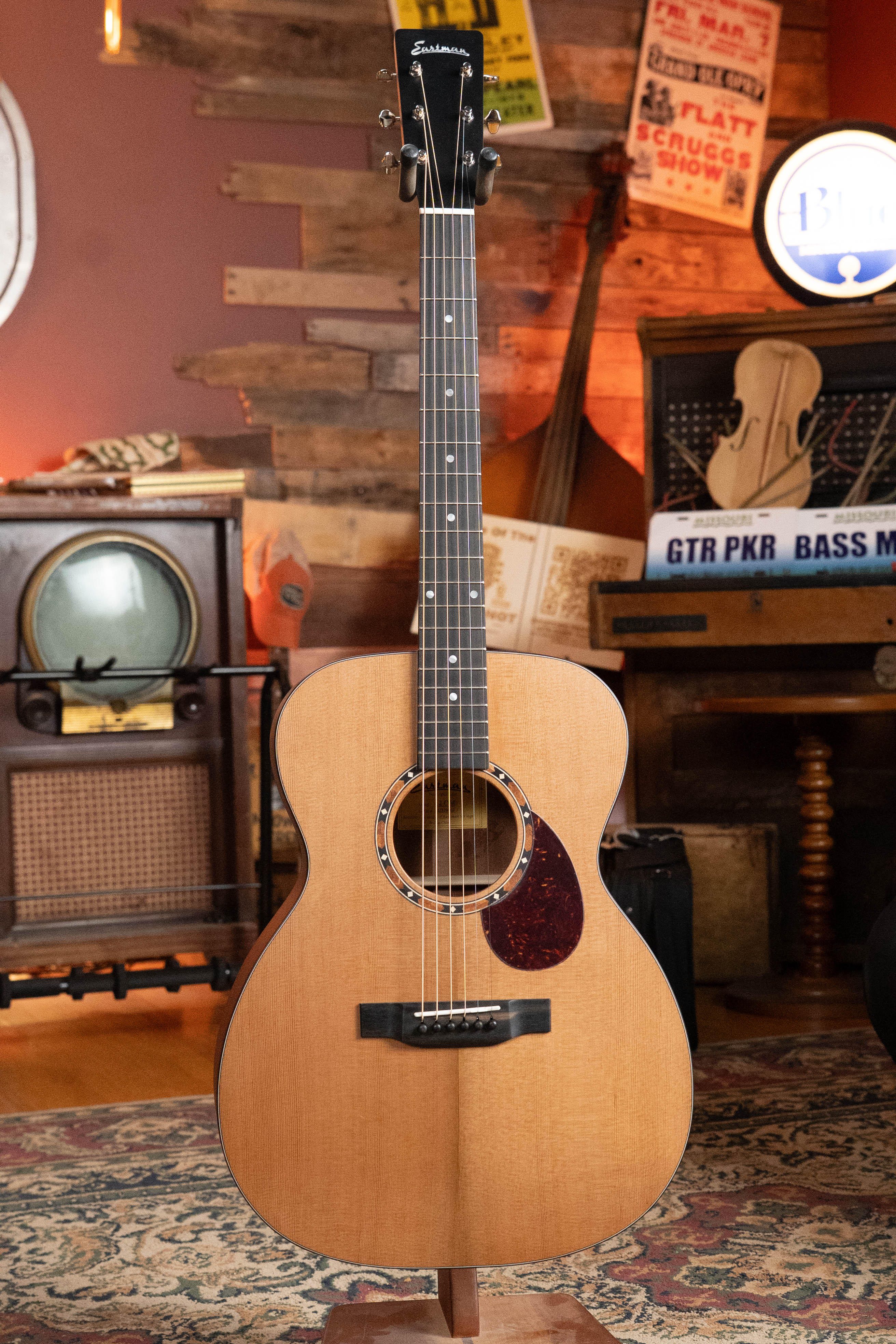 An Eastman Guitars E2OM Cedar/Sapele Orchestra Model Acoustic Guitar #9002 with a natural finish stands upright in a cozy vintage room filled with musical instruments, retro signs, and an old radio.