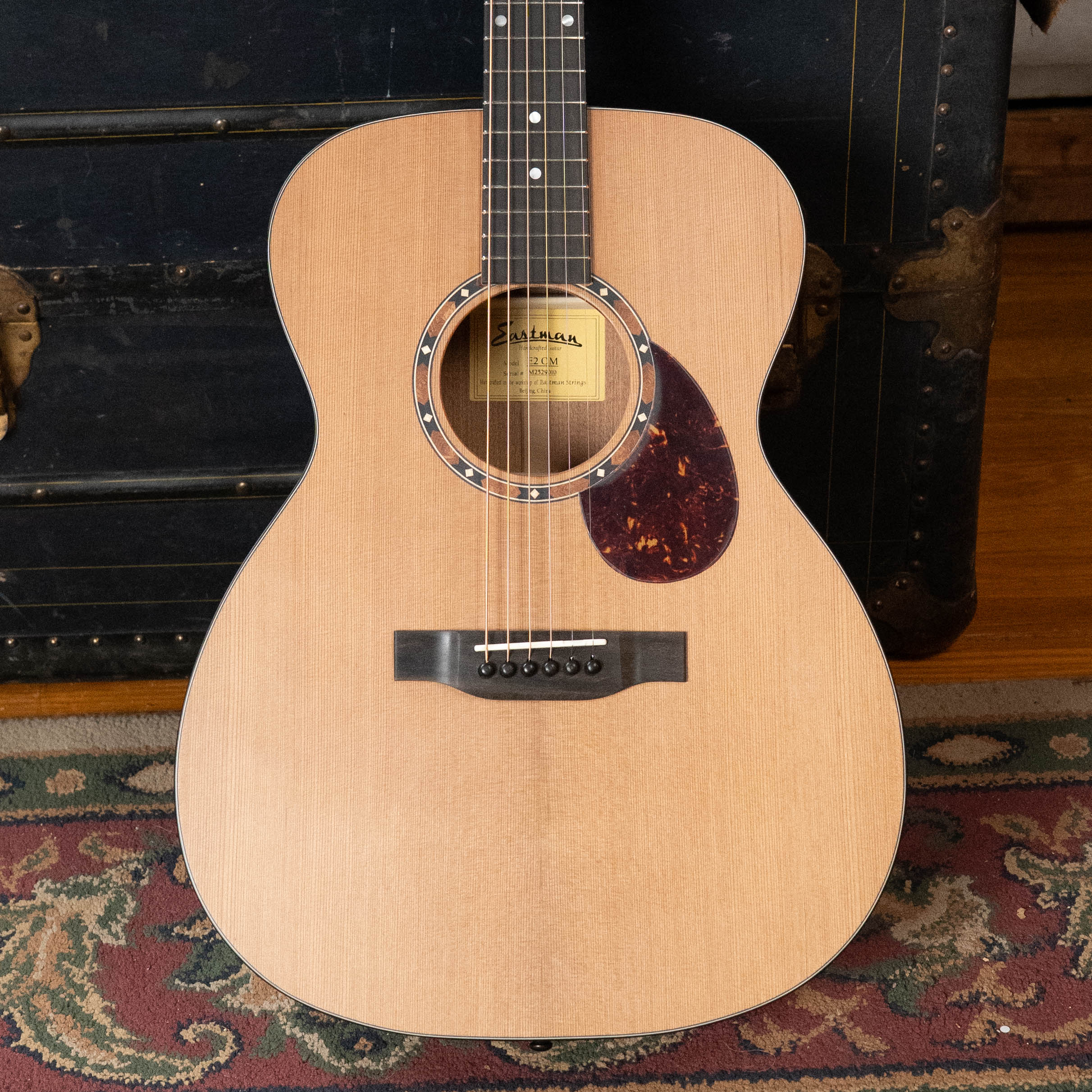 An Eastman Guitars E2OM Cedar/Sapele Orchestra Model Acoustic Guitar #9010 rests upright against a vintage trunk on a patterned rug, featuring a cedar top and classic styling.