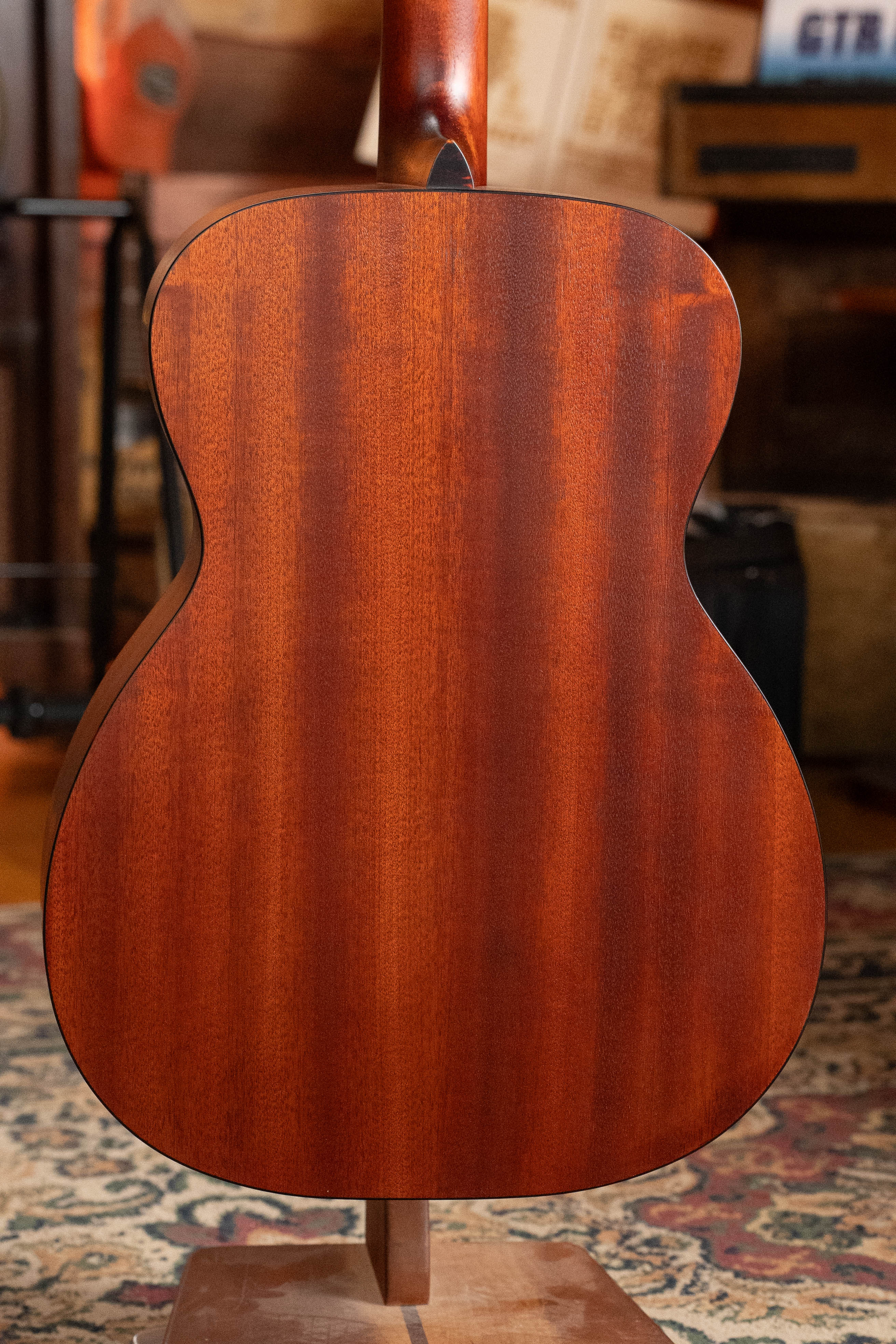A close-up of the back of the Eastman Guitars E2OM Cedar/Sapele Orchestra Model Acoustic Guitar #9010, with its rich, reddish-brown wood finish, standing on a wooden stand before a patterned rug and blurred musical instruments.