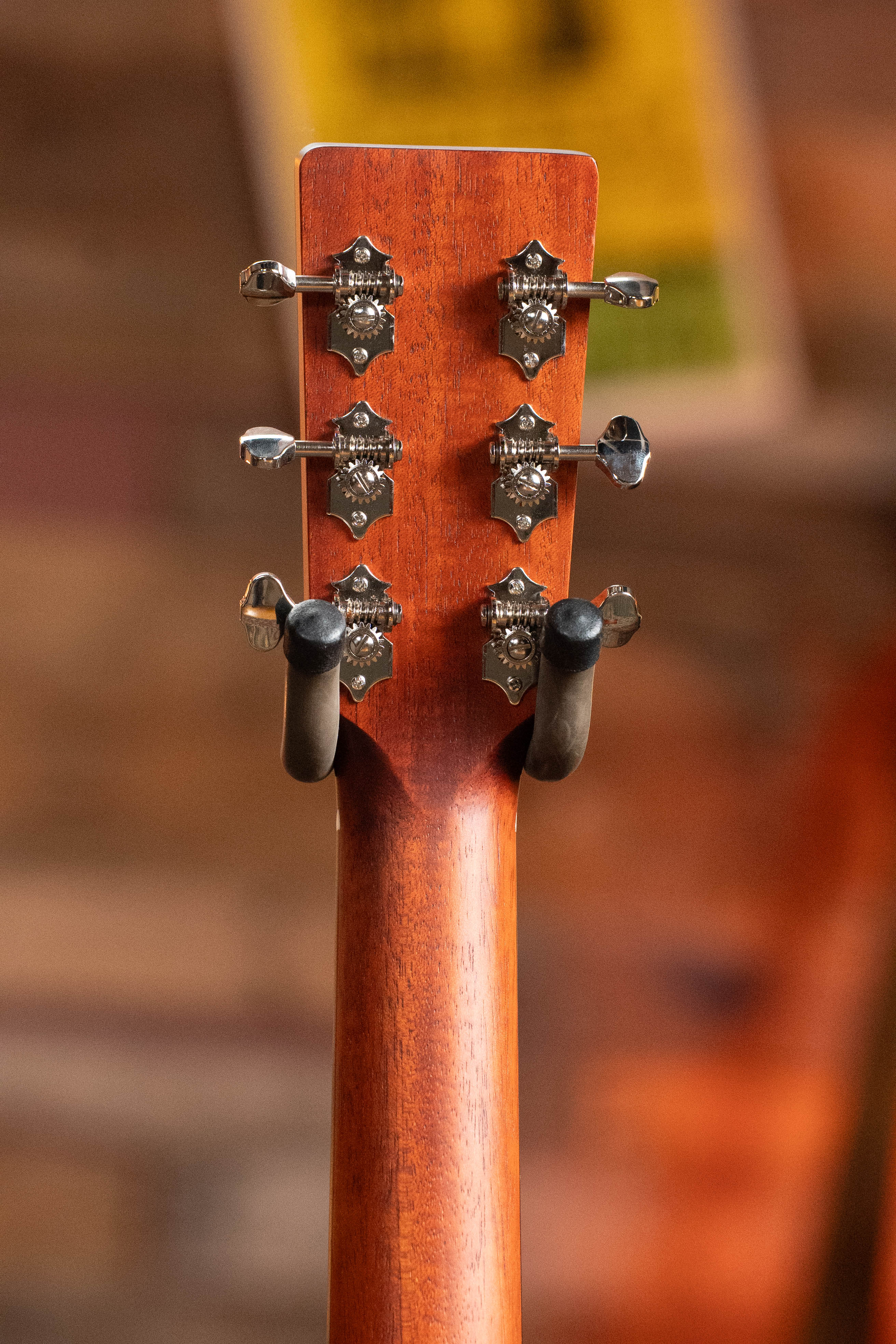 The back of an Eastman Guitars E2OM Cedar/Sapele Orchestra Model Acoustic Guitar #9010 headstock features six metal tuning pegs and a reddish-brown finish, displayed on a wall hanger with a softly blurred background.