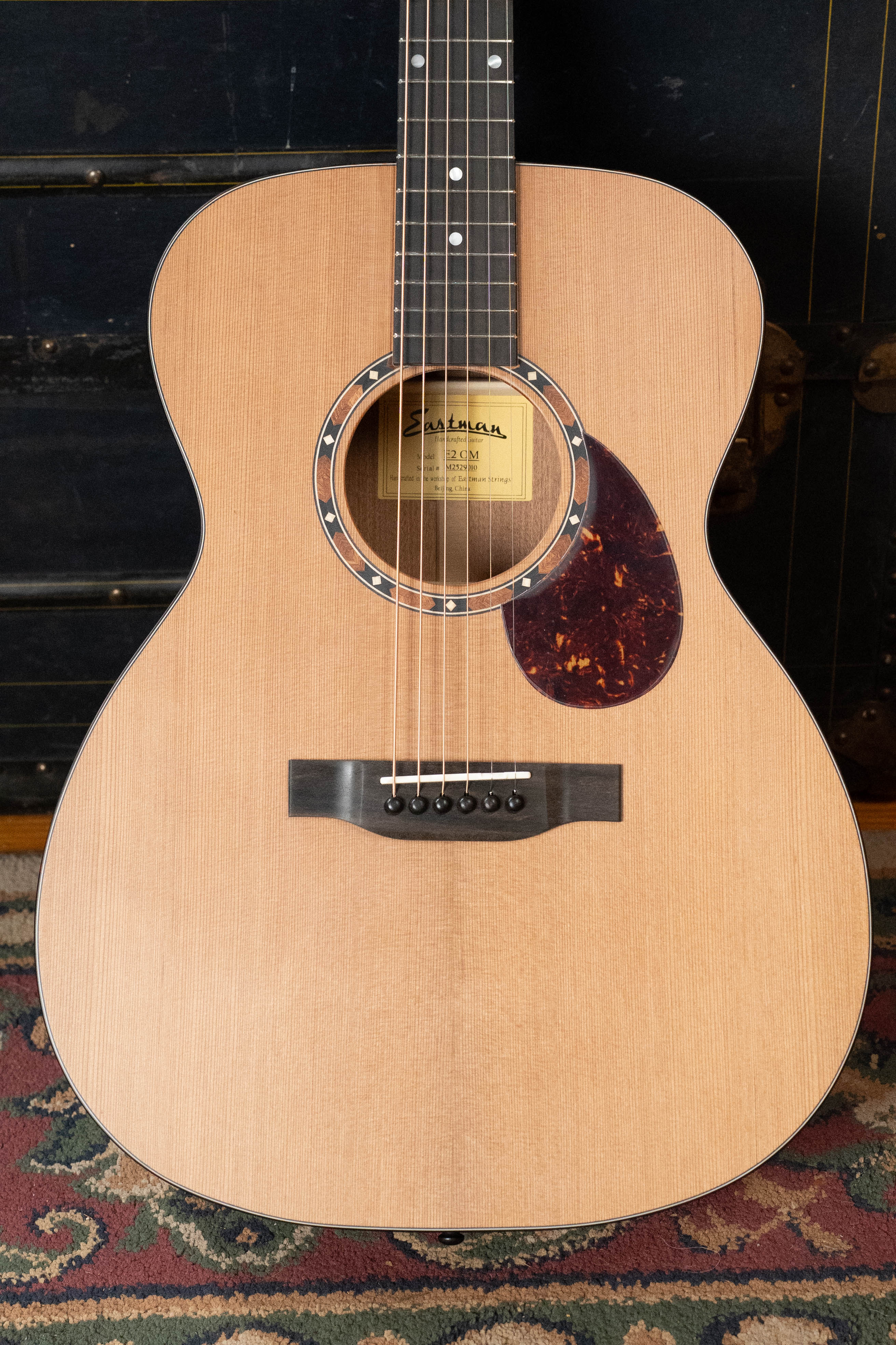 An Eastman Guitars E2OM Cedar/Sapele Orchestra Model Acoustic Guitar #9010 with a natural finish and tortoiseshell-style pickguard rests on a patterned rug with a black trunk in the background—ideal for fingerstyle playing.