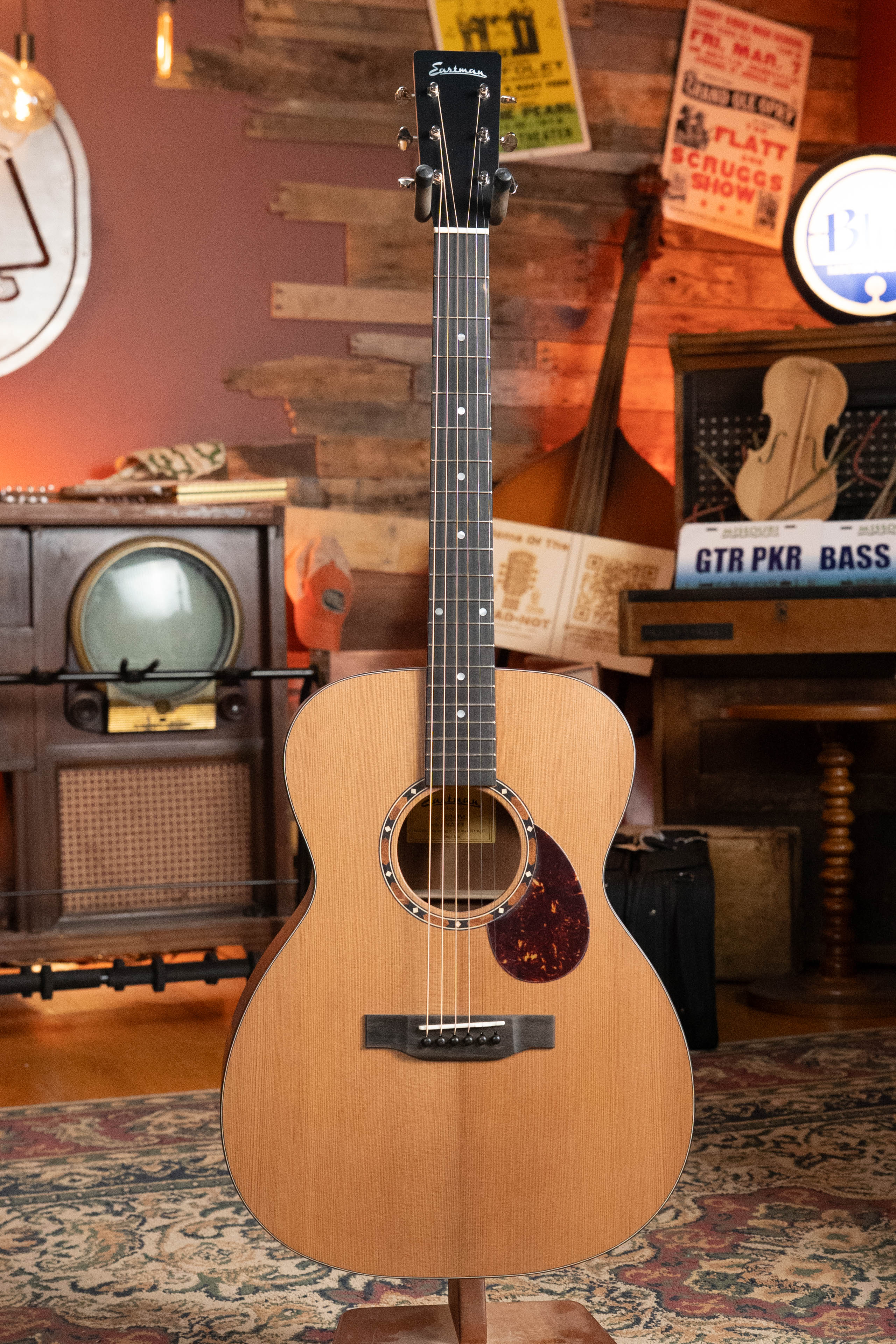 An Eastman E2OM Cedar/Sapele Orchestra Model Acoustic Guitar by Eastman Guitars sits on a stand in a cozy, vintage room filled with musical instruments, an old radio, posters, and warm lighting. Ideal for fingerstyle playing.