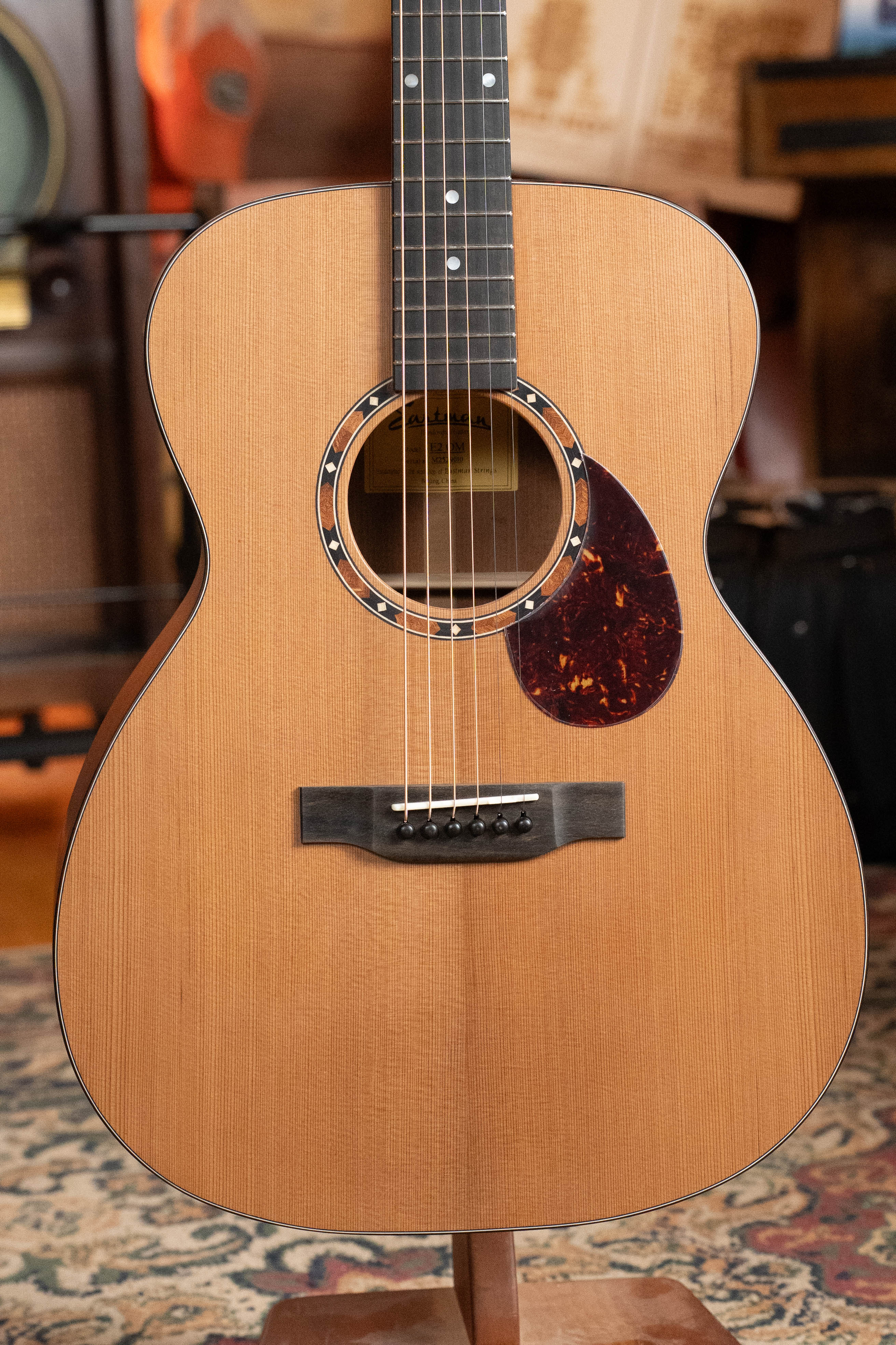 The Eastman Guitars E2OM Cedar/Sapele Orchestra Model Acoustic Guitar #9010 features a natural finish, black pickguard, and six strings, shown upright on a stand with musical gear—a great choice for fingerstyle players.