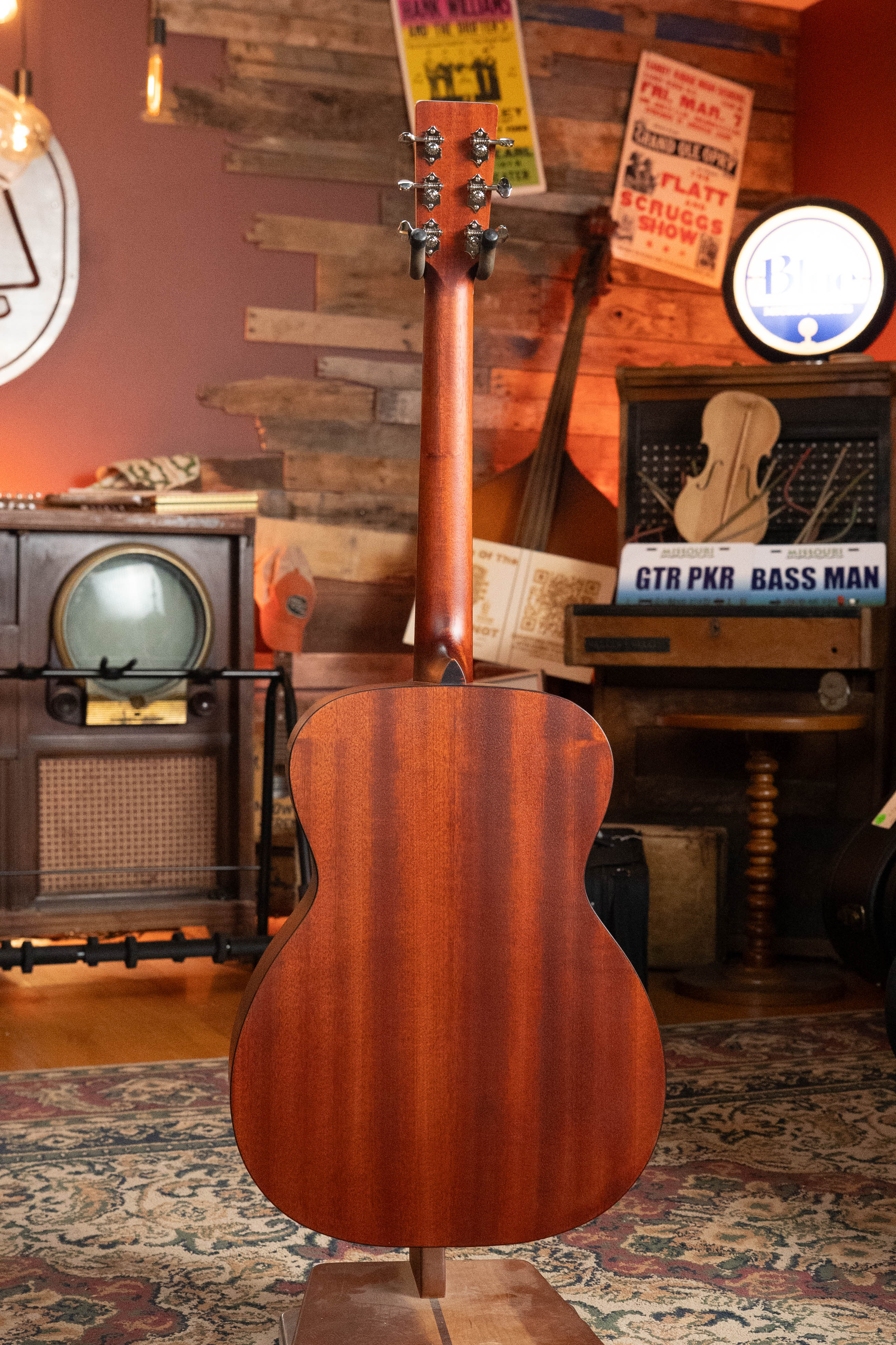The Eastman E2OM Cedar/Sapele Orchestra Model Acoustic Guitar #9010 by Eastman Guitars is showcased with its back facing forward in a cozy, vintage-style music room—ideal for fingerstyle guitar enthusiasts.