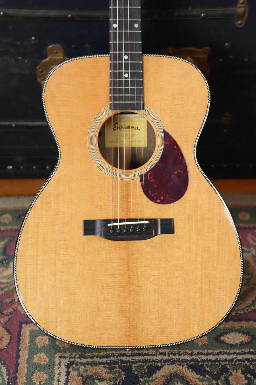 A close-up of the Eastman Guitars E3OM-DLX Sitka/Ovangkol Acoustic Guitar w/ Fishman Pickup #9222 showcases its solid Sitka spruce top, black pickguard, and six strings on a patterned rug with a trunk featuring brass hardware in the background.