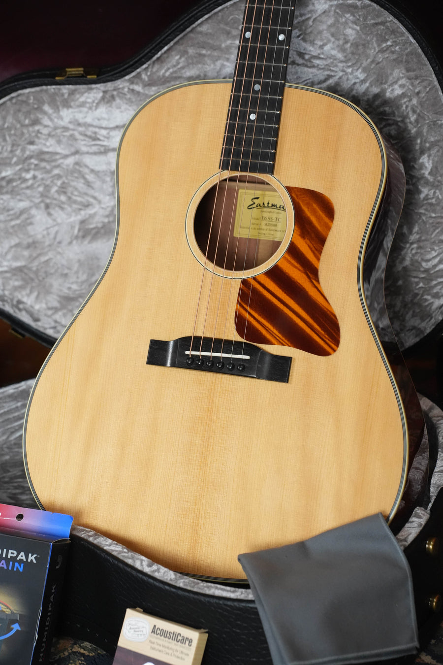 A close-up of the Eastman Guitars E6SS-TC Thermo-Cured Slope Shoulder Dreadnought Acoustic Guitar #1116 with a natural finish sits in an open case, with included accessories like a cleaning cloth and string pack visible in front.
