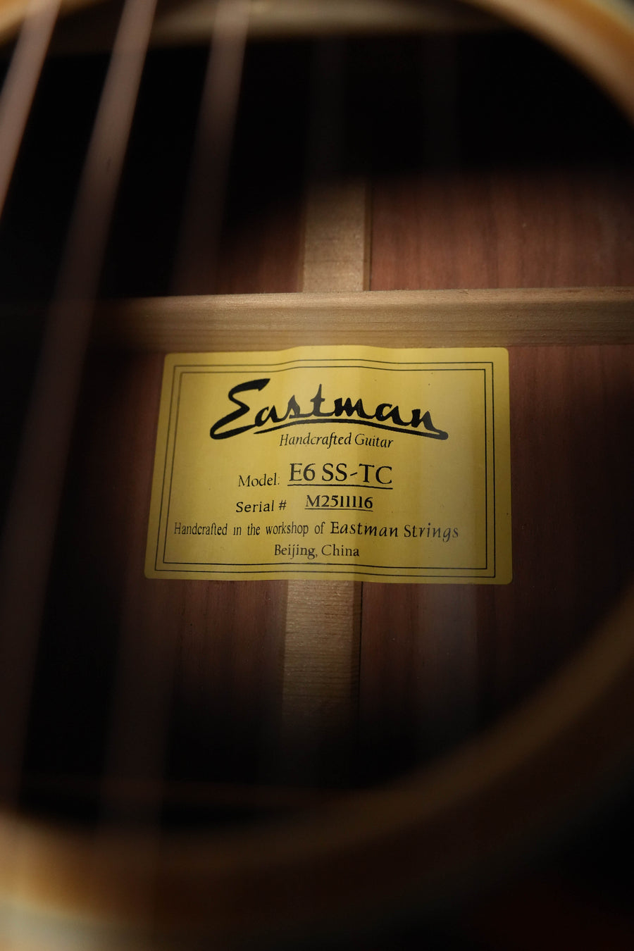 Close-up through the strings of an Eastman E6SS-TC Thermo-Cured Slope Shoulder Dreadnought Acoustic Guitar #1116 by Eastman Guitars shows its yellow label with model and serial number, crafted in Beijing, China.