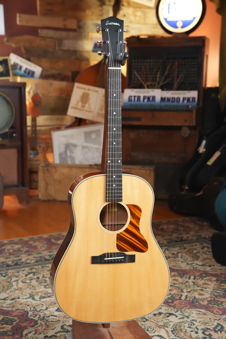 An Eastman Guitars E6SS-TC Thermo-Cured Slope Shoulder Dreadnought Acoustic Guitar #1116 with a natural finish stands upright in a cozy vintage room featuring warm lighting, wooden décor, a patterned rug, and musical equipment.