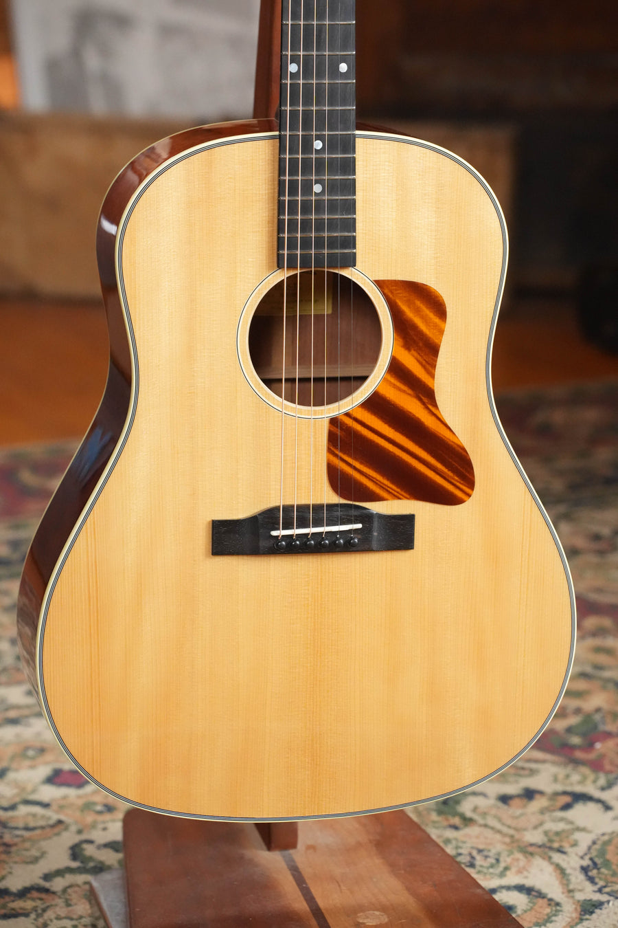 A close-up of the Eastman Guitars E6SS-TC Thermo-Cured Slope Shoulder Dreadnought Acoustic Guitar #1116, featuring a natural wood finish, black fretboard, and striped wood pickguard on a wooden stand over a patterned rug.