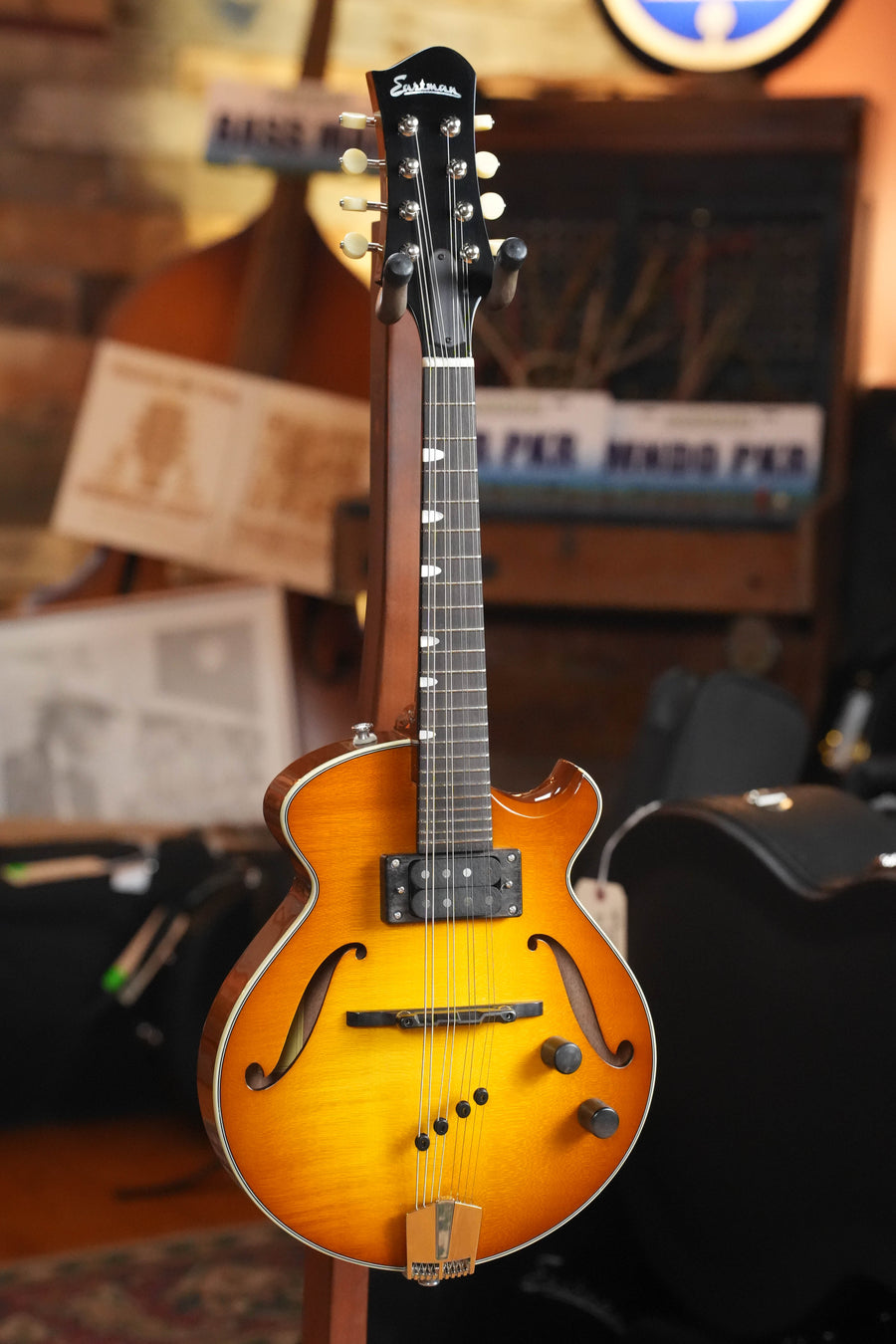 An Eastman Guitars ER-M-GB Goldburst El Rey Mandolin #0211 with F-holes and two pickups is displayed on a stand indoors, surrounded by musical equipment and blurred sheet music in the background.