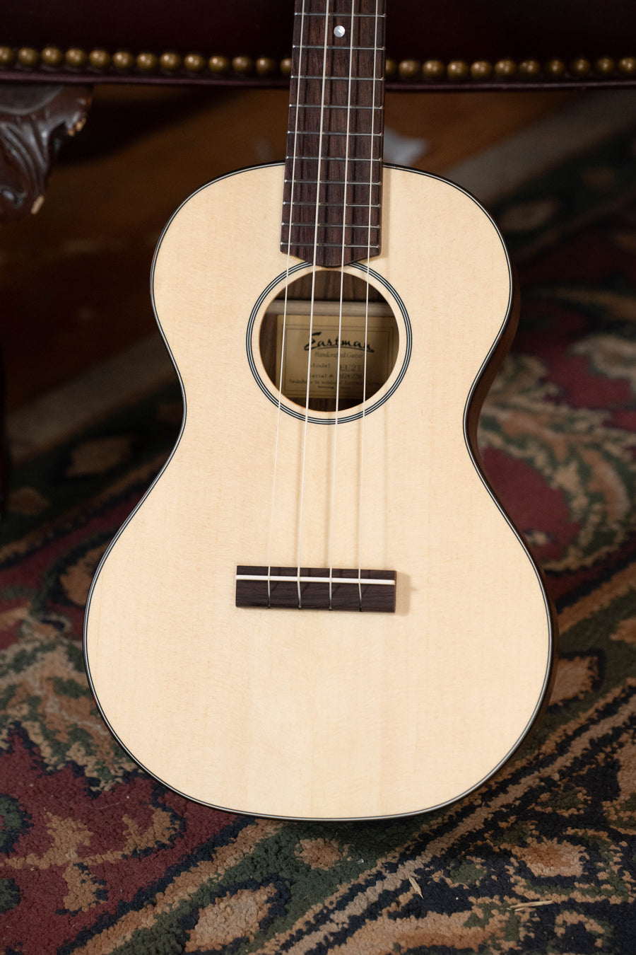 A close-up of an Eastman Guitars EU2-T Spruce/Ovangkol Tenor Ukulele (#7761) rests on a patterned rug, with a brown leather chair partially visible in the background.