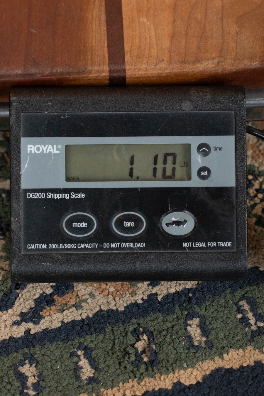 A digital shipping scale reads 1.10 lbs with several buttons, sitting on patterned carpet and supporting part of an Eastman Guitars EU2-T Spruce/Ovangkol Tenor Ukulele #7761.