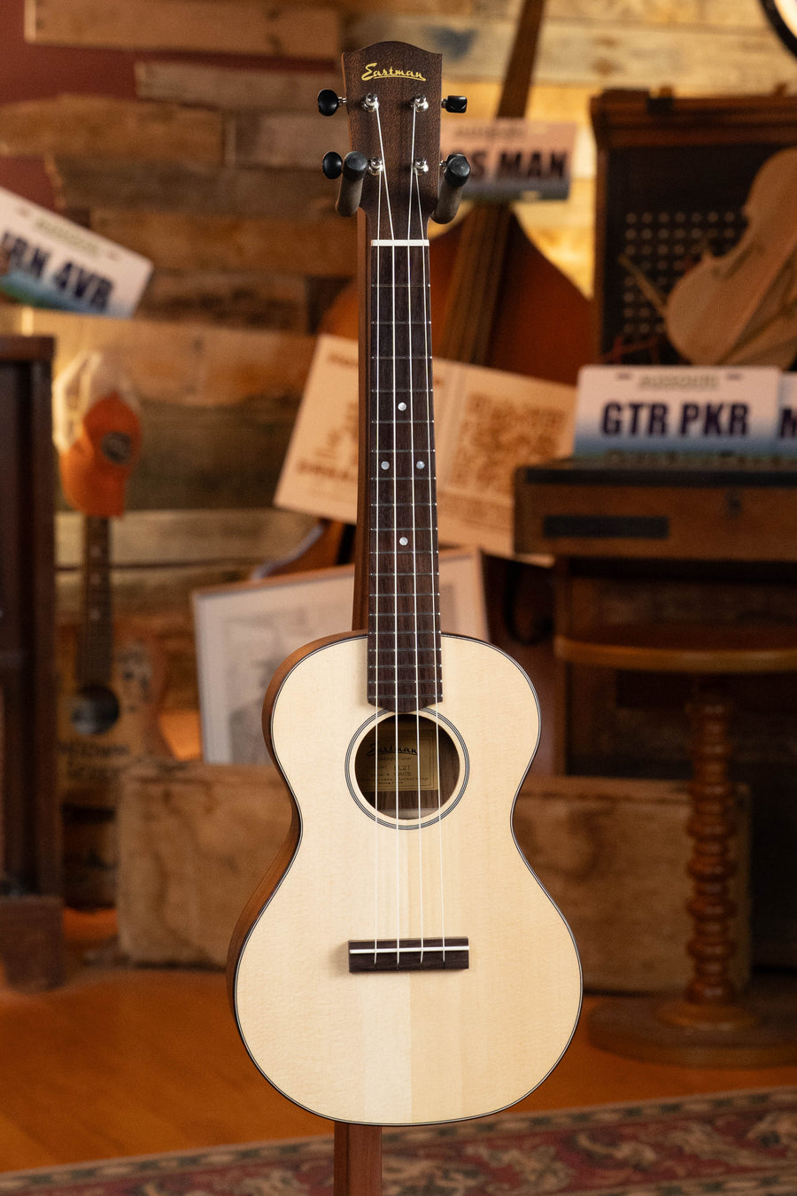 The Eastman Guitars EU2-T Spruce/Ovangkol Tenor Ukulele #7761, with a light body and dark headstock and fretboard, stands upright in a rustic room featuring wooden decor and vintage instruments in the background.