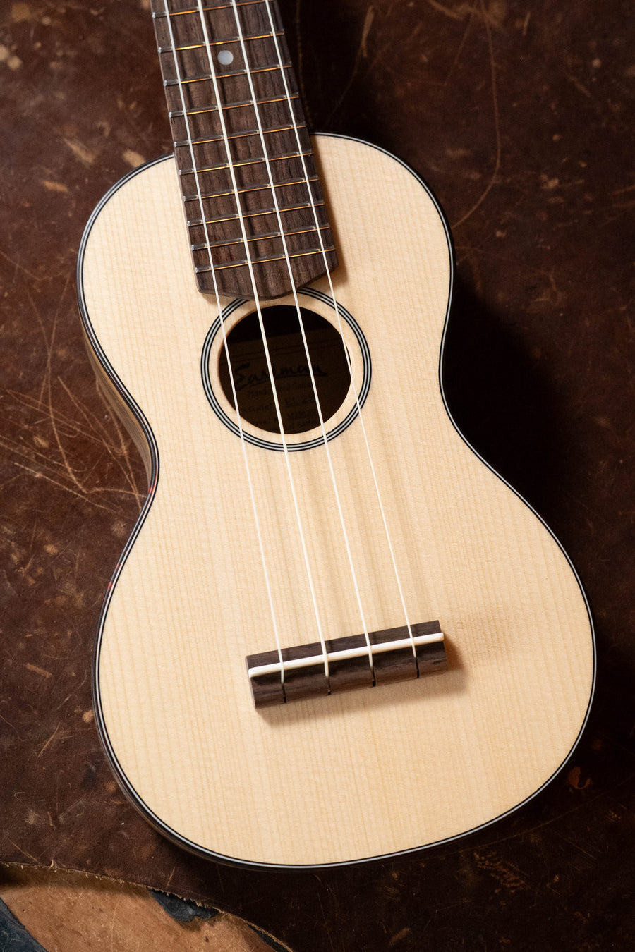 A close-up of the Eastman Guitars EU2-s Sitka/Rosewood Soprano Ukulele (#2071) with four strings, showing its sound hole and fretboard, resting on a dark brown textured surface.