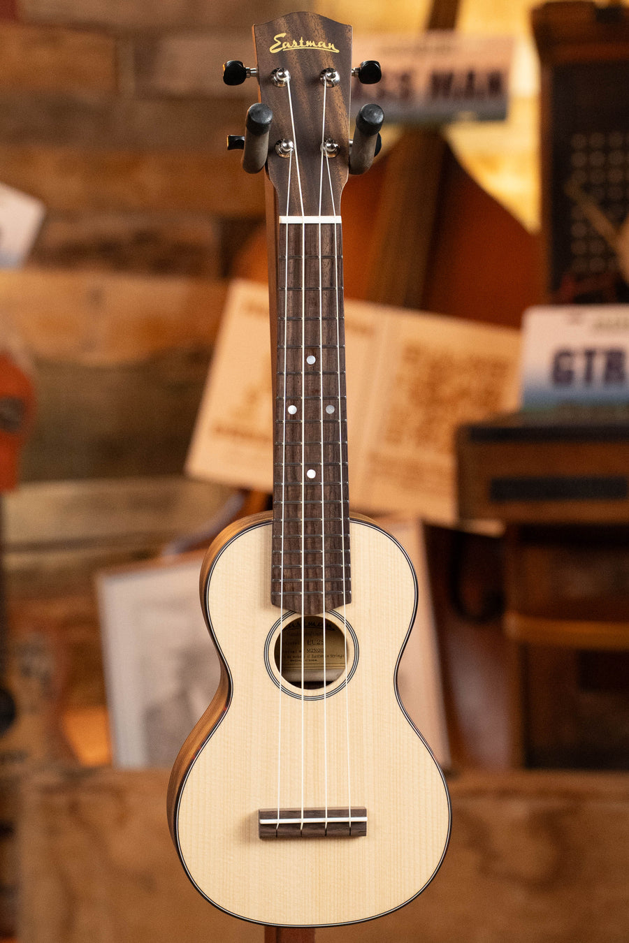The Eastman Guitars EU2-s Sitka/Rosewood Soprano Ukulele #2071, with a light solid Sitka wood body and dark fretboard, stands upright against a blurred backdrop of sheet music and other wooden instruments.