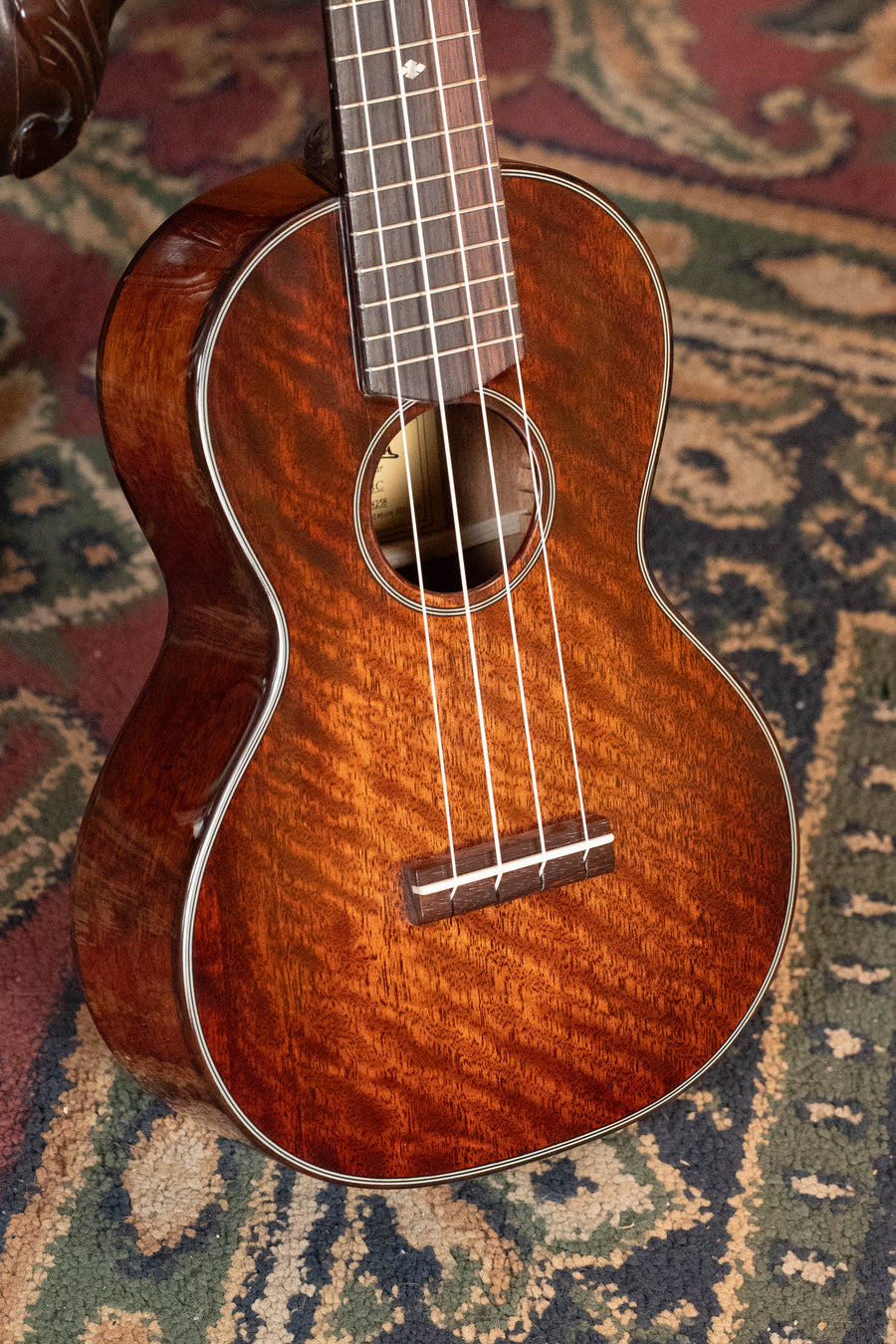 A close-up of the Eastman Guitars EU3-C All Mahogany Concert Ukulele #8258 shows its rich, glossy finish, visible strings, fretboard, and curvy body as it rests on a patterned carpet.