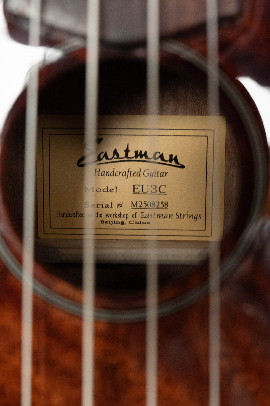 Close-up of a concert ukulele’s soundhole reveals a label reading Eastman Guitars, Model: EU3-C All Mahogany Concert Ukulele #8258, showcasing the craftsmanship of every Eastman instrument made in Beijing, China.