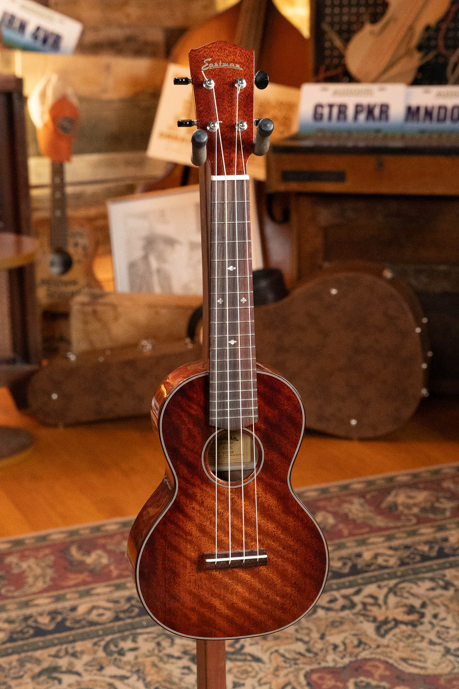 The Eastman EU3-C All Mahogany Concert Ukulele by Eastman Guitars stands upright on a stand in a cozy room with wooden floors, a patterned rug, and various musical instruments and cases in the background.