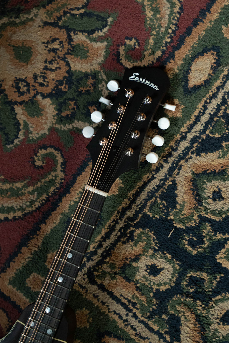 A close-up of the headstock of an Eastman Guitars MD304 Spruce/Maple A-Style Oval Hole Mandolin #4581 with white tuning pegs, resting on a patterned green, red, and beige carpet.