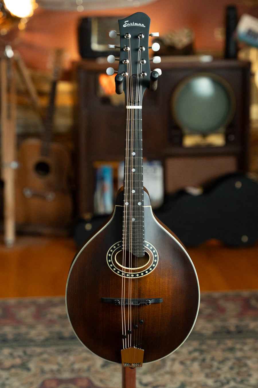 An Eastman Guitars MD304 Spruce/Maple A-Style Oval Hole Mandolin #4581 with a sunburst finish stands upright in a warmly lit room, surrounded by vintage musical instruments and an old television set.