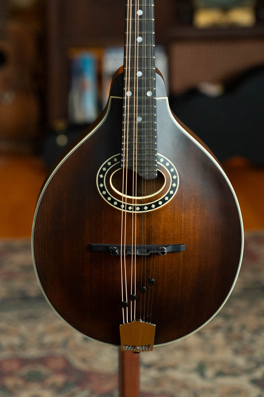 A close-up of the Eastman MD304 Spruce/Maple A-Style Oval Hole Mandolin (#4581) by Eastman Guitars, featuring a dark brown sunburst finish and decorative inlay, resting on a stand indoors with a patterned rug in the background.