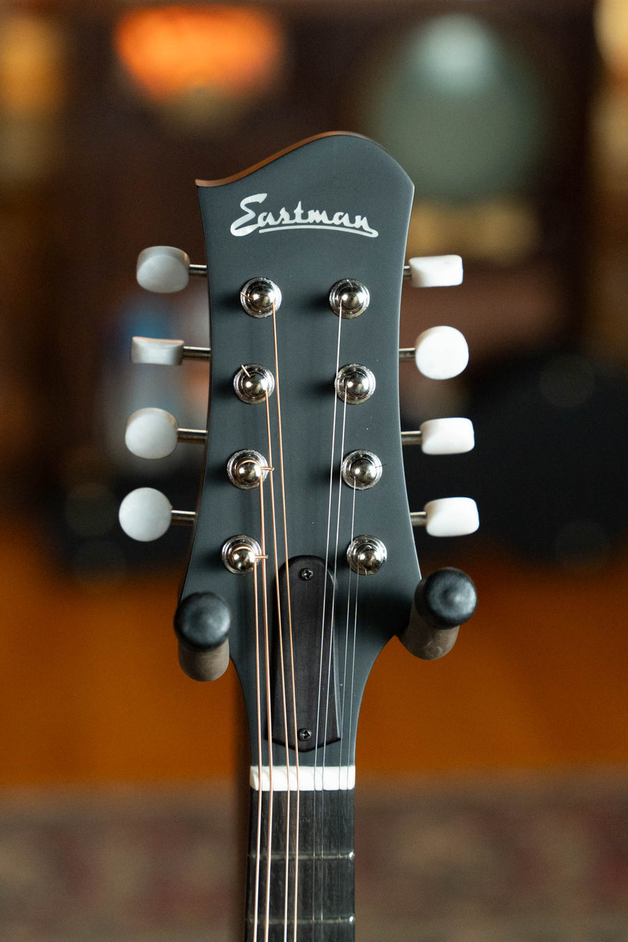 Close-up of the Eastman Guitars MD304 Spruce/Maple A-Style Oval Hole Mandolin #4581 headstock in black, featuring silver tuning pegs and white buttons, sharply in focus against a blurred background.