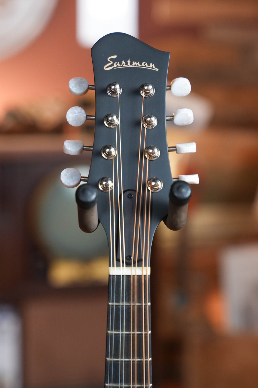 Close-up of the headstock of an Eastman Guitars MD305L Left-Handed Spruce/Maple A-Style Hand-Carved Mandolin #4951 with six tuning pegs and strings, resting on a hanger against a blurred, warm-toned background.