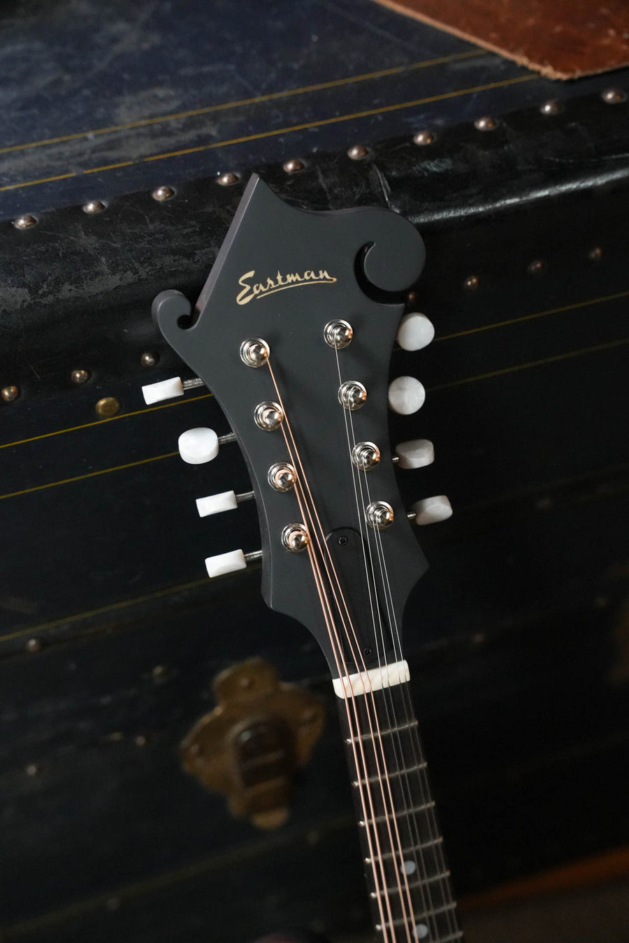 Close-up of the headstock of an Eastman Guitars MD314 Spruce/Maple F-Style Oval Hole Mandolin #3716, with tuning pegs and strings, leaning on a dark vintage trunk with brass hardware.