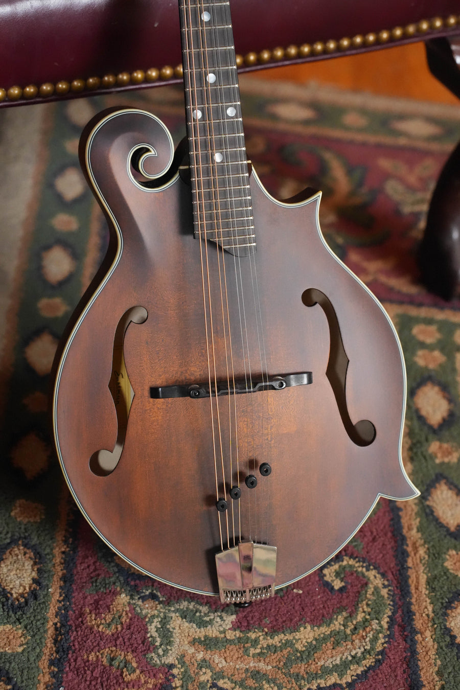 A close-up of the Eastman Guitars MD315 F-Style Hand-Carved Mandolin #0107 features its ornate scroll and f-holes, resting on a patterned carpet with a leather chair in the background, highlighting its professional-level tone.