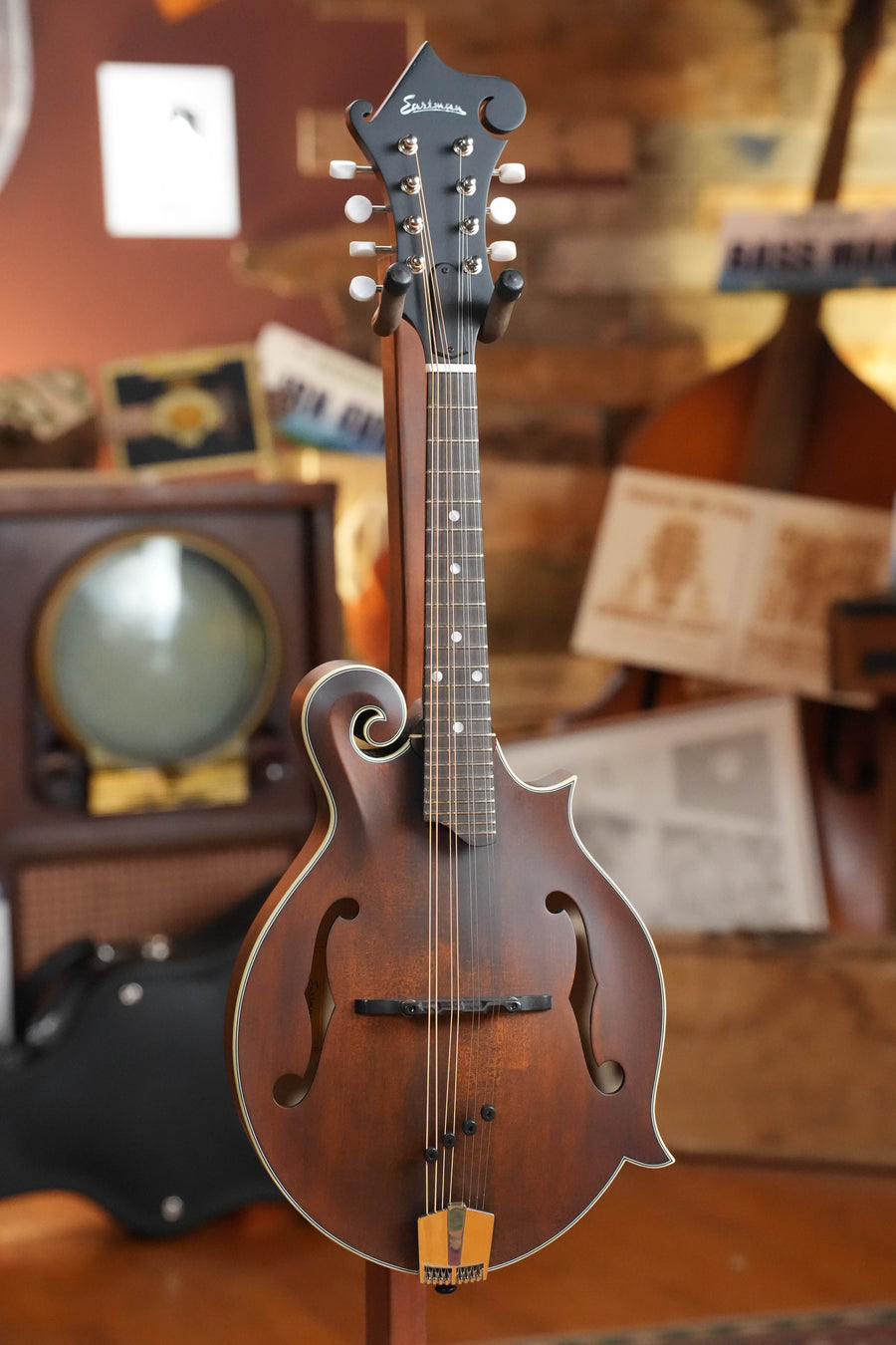 The Eastman Guitars MD315 F-Style Hand-Carved Mandolin #0107 stands upright on a display, set against a wooden backdrop with vintage music gear, highlighting its pro-level tone and timeless design.