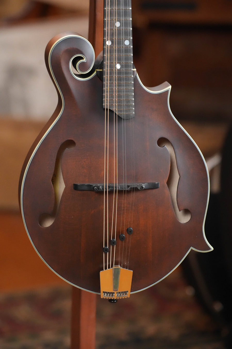 A close-up of the Eastman MD315 F-Style Hand-Carved Mandolin #0329 by Eastman Guitars, featuring ornate scrollwork, f-holes, and a gold tailpiece, stands upright indoors—ready for professional-level tone.