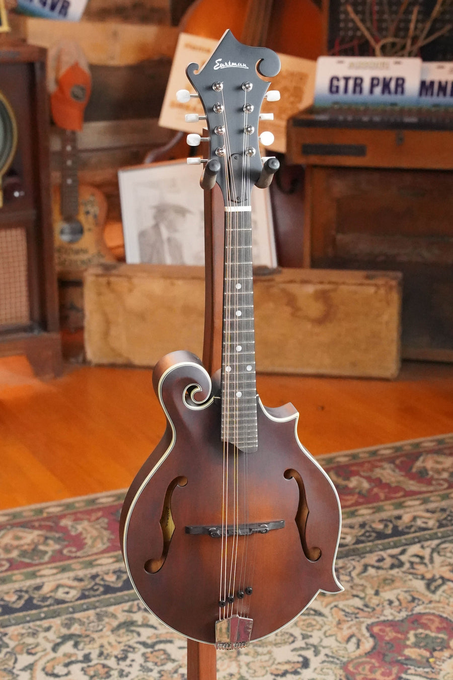An Eastman Guitars MD315 F-Style Hand-Carved Mandolin #2483 with a scroll design is displayed on a stand in a cozy room with wooden floors, vintage decor, and other instruments—reflecting its professional-level tone.