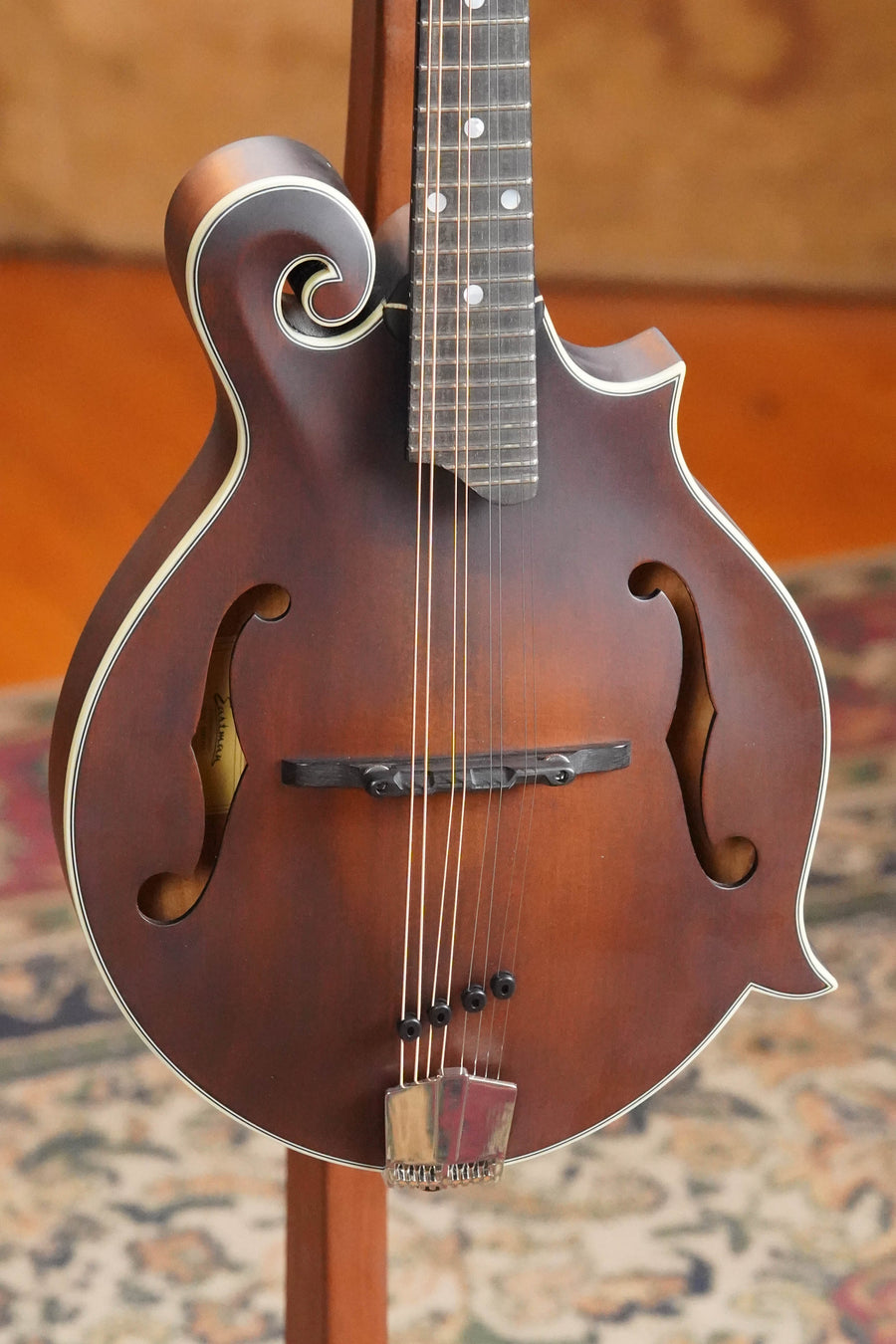 A close-up of an Eastman Guitars MD315 F-Style Hand-Carved Mandolin #2483 showcases its decorative curves and dual f-shaped sound holes on a stand, with a patterned rug and wooden floor highlighting its expert craftsmanship.