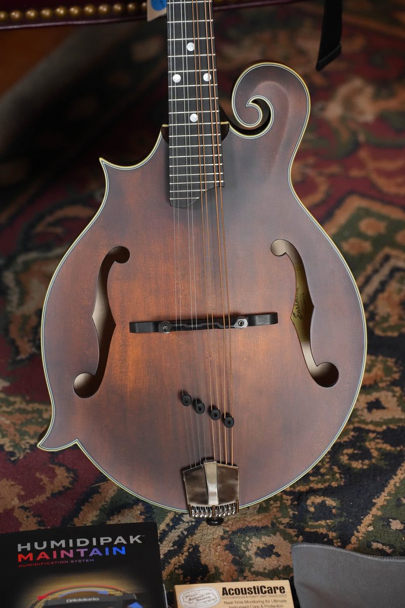 A close-up of the Eastman Guitars MD315L Left-Handed Spruce/Maple F-Style Hand-Carved Mandolin #3427, featuring an ornate headstock and f-holes, rests on a patterned rug beside maintenance items including Humidipak Maintain.