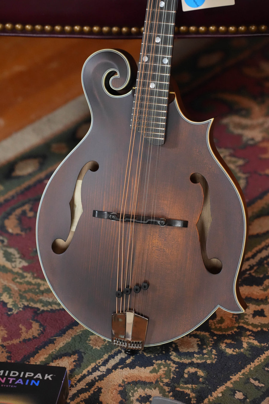 A close-up of an Eastman Guitars MD315 Spruce/Maple F-Style Hand-Carved Mandolin #5783, featuring ornate curves and f-holes, resting on a patterned rug with a book visible in the foreground.