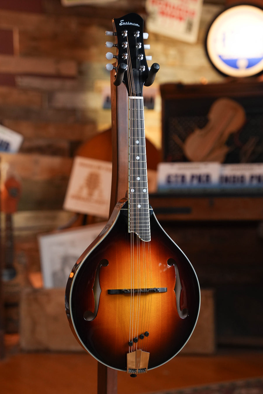 The Eastman Guitars MD505-CS Spruce/Maple Classic Sunburst A-Style Mandolin #3162, featuring a teardrop shape, is displayed on a stand in a warmly lit room with music-themed decor in the background.