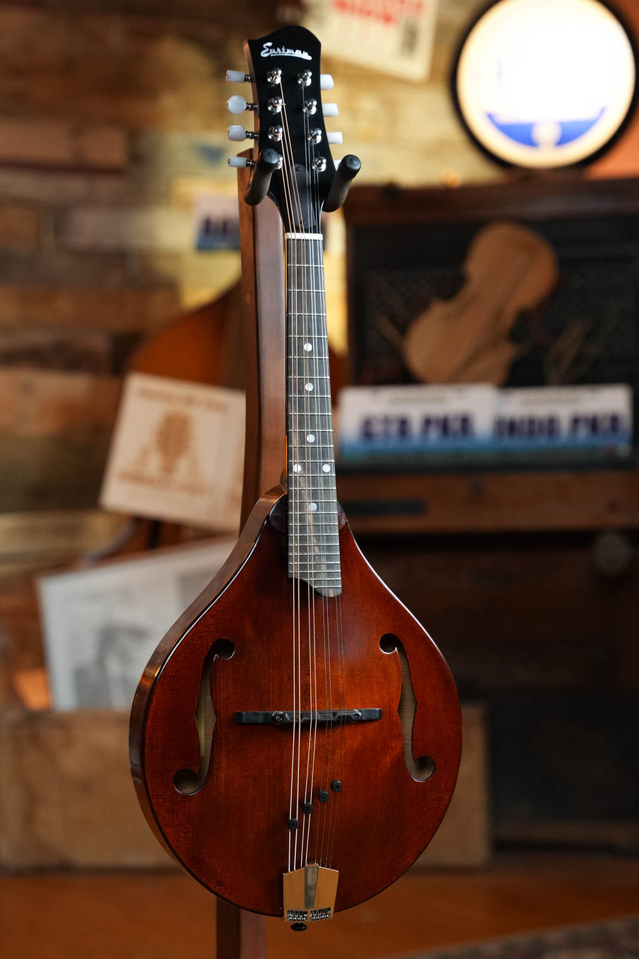 An Eastman Guitars MD505CC/TV Comfort Contour True Tone Vintage Series A Style Mandolin (#2859) stands upright on a stand, with its curved body and F-holes highlighted against a blurred wooden and postered background.