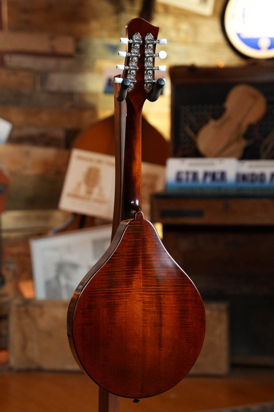 The Eastman Guitars MD505CC/TV Comfort Contour True Tone Vintage Series A Style Mandolin #2859 is shown from the back with its glossy brown finish, upright in a warmly lit room amid musical instruments and sheet music.