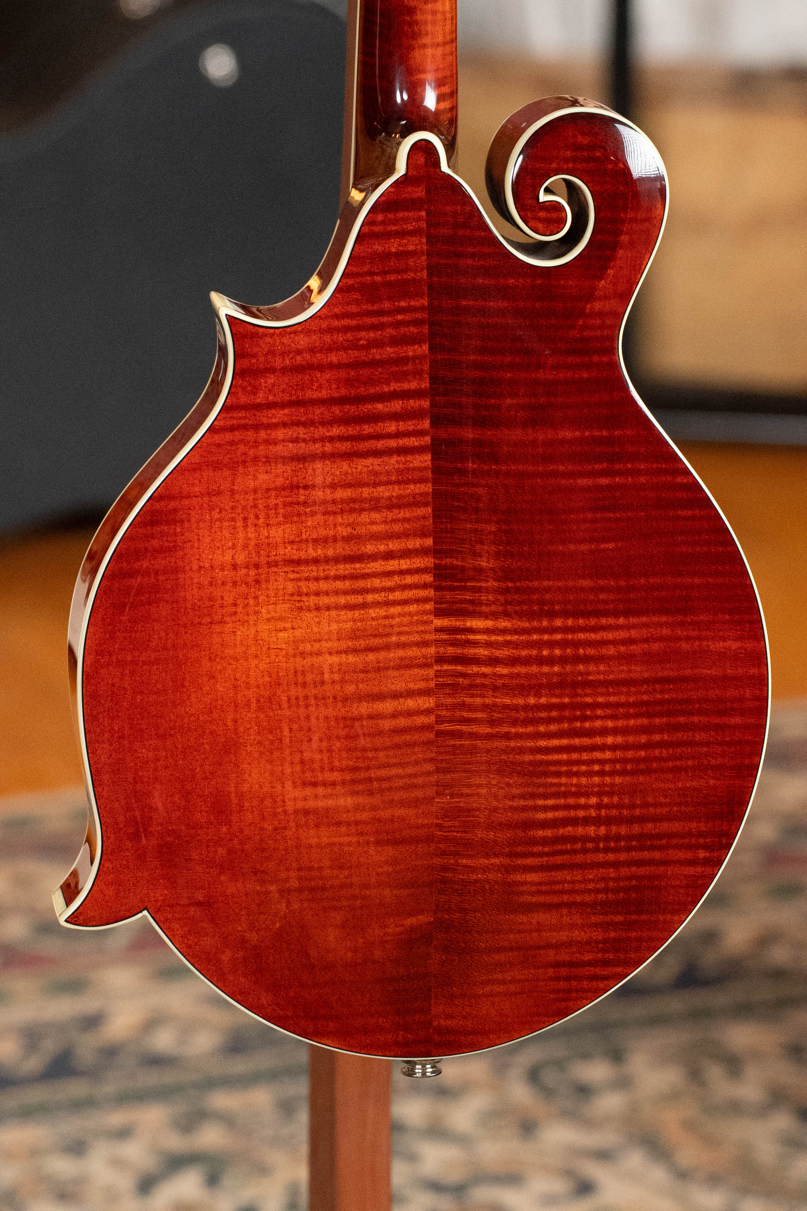 Close-up of the back of the Eastman Guitars MD615 Spruce/Maple F-Style Mandolin w/K&K Pickup #3918, featuring a glossy reddish-brown finish, cream binding, and ornate scrollwork against a blurred indoor background.
