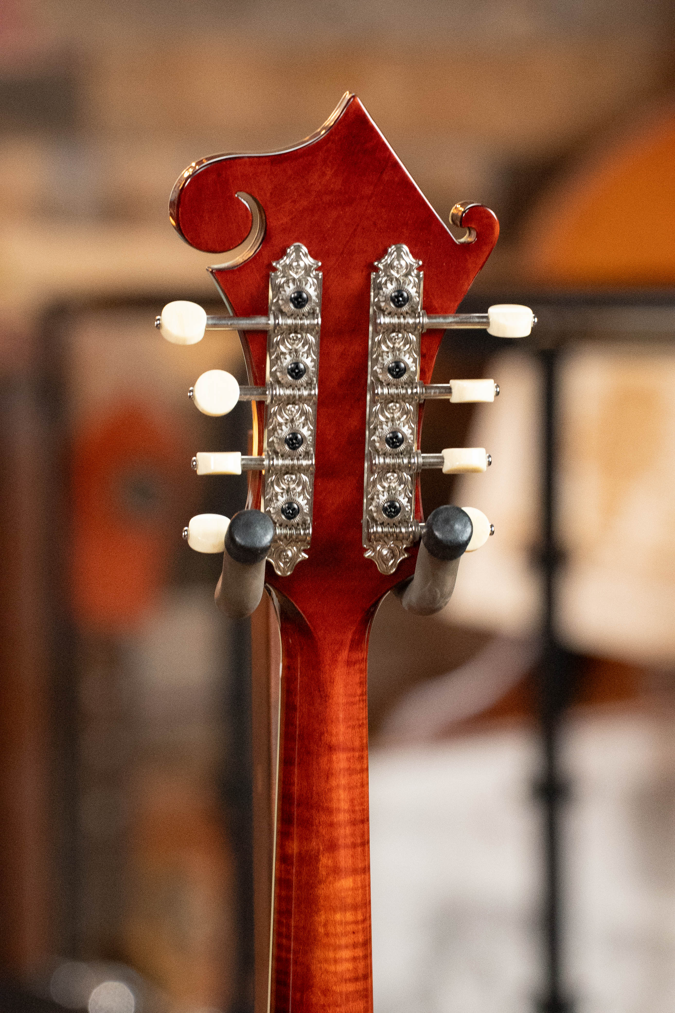 Close-up of the Eastman Guitars MD615 Spruce/Maple F-Style Mandolin w/K&K Pickup (#3918) headstock, showing ornate metal tuning pegs with white knobs and a reddish wood finish, set against a blurred background.