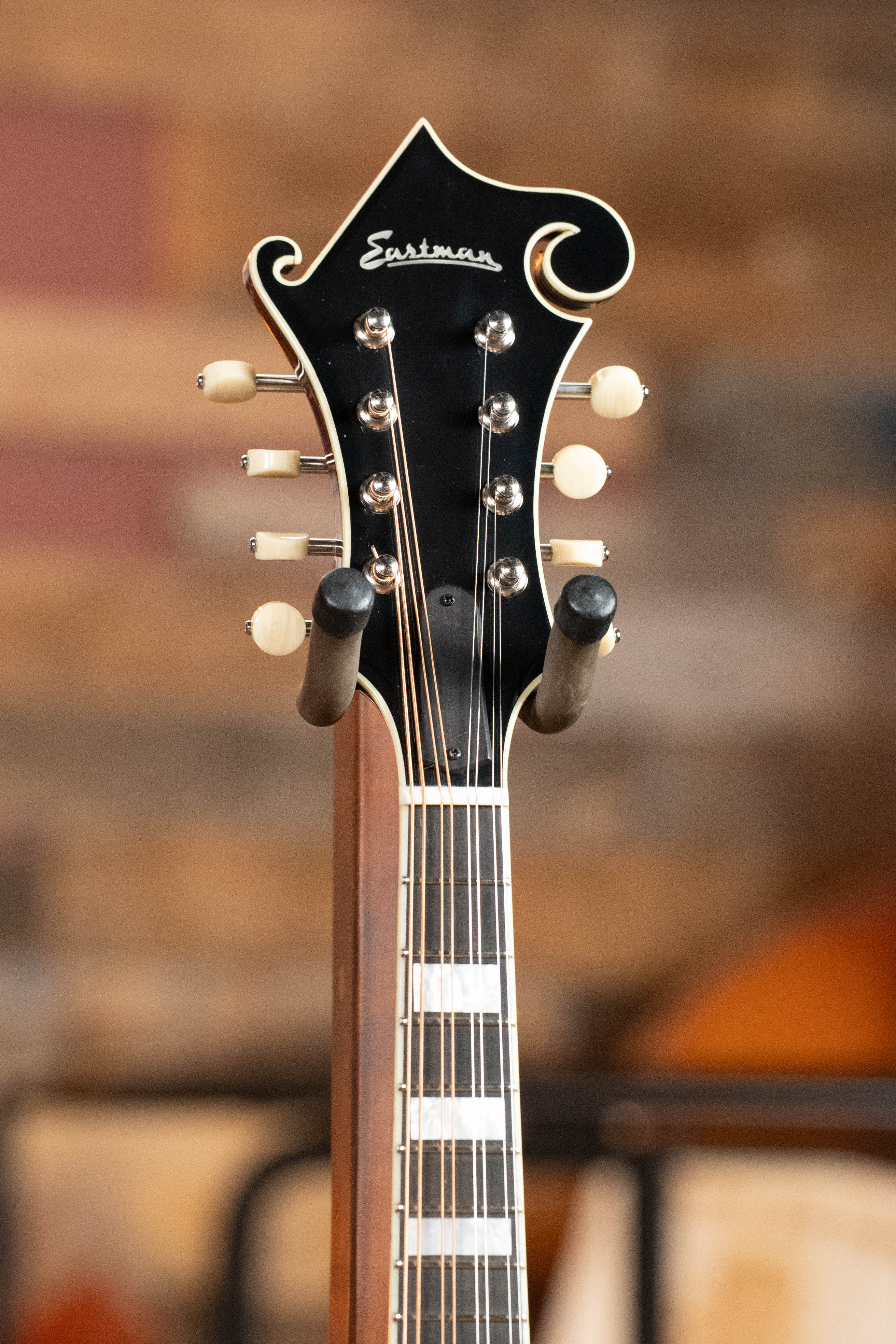 Close-up of the headstock and fretboard of an Eastman Guitars MD615 Spruce/Maple F-Style Mandolin w/K&K Pickup #3918, featuring white tuning pegs and a black finish against a softly blurred, warm-toned background.
