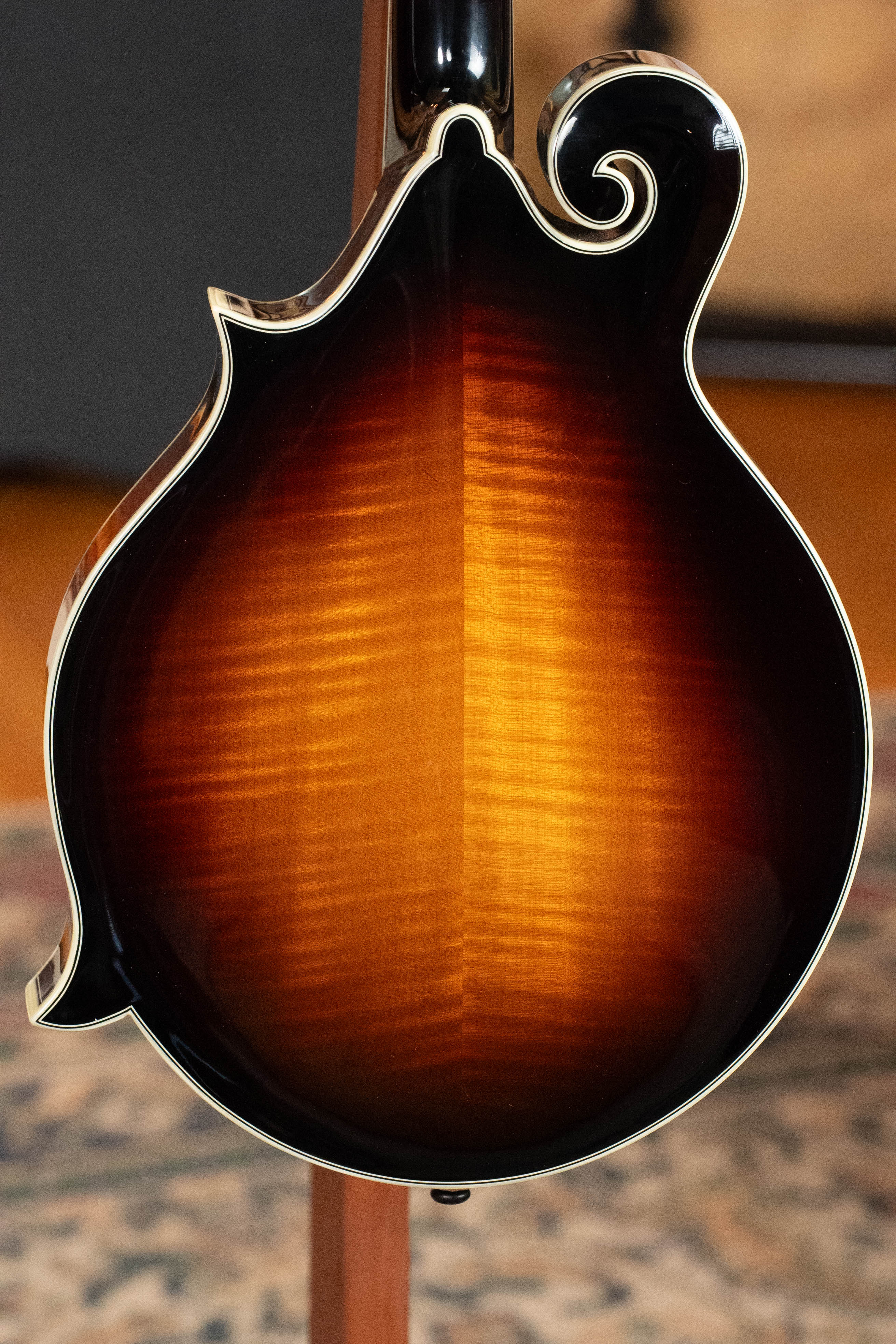 The image shows the back of an Eastman Guitars MD815-SB Adirondack/Flamed Maple Sunburst F-Style Mandolin #3355, displaying a glossy amber-to-dark brown sunburst finish with a decorative curl on the upper right, resting on a stand.
